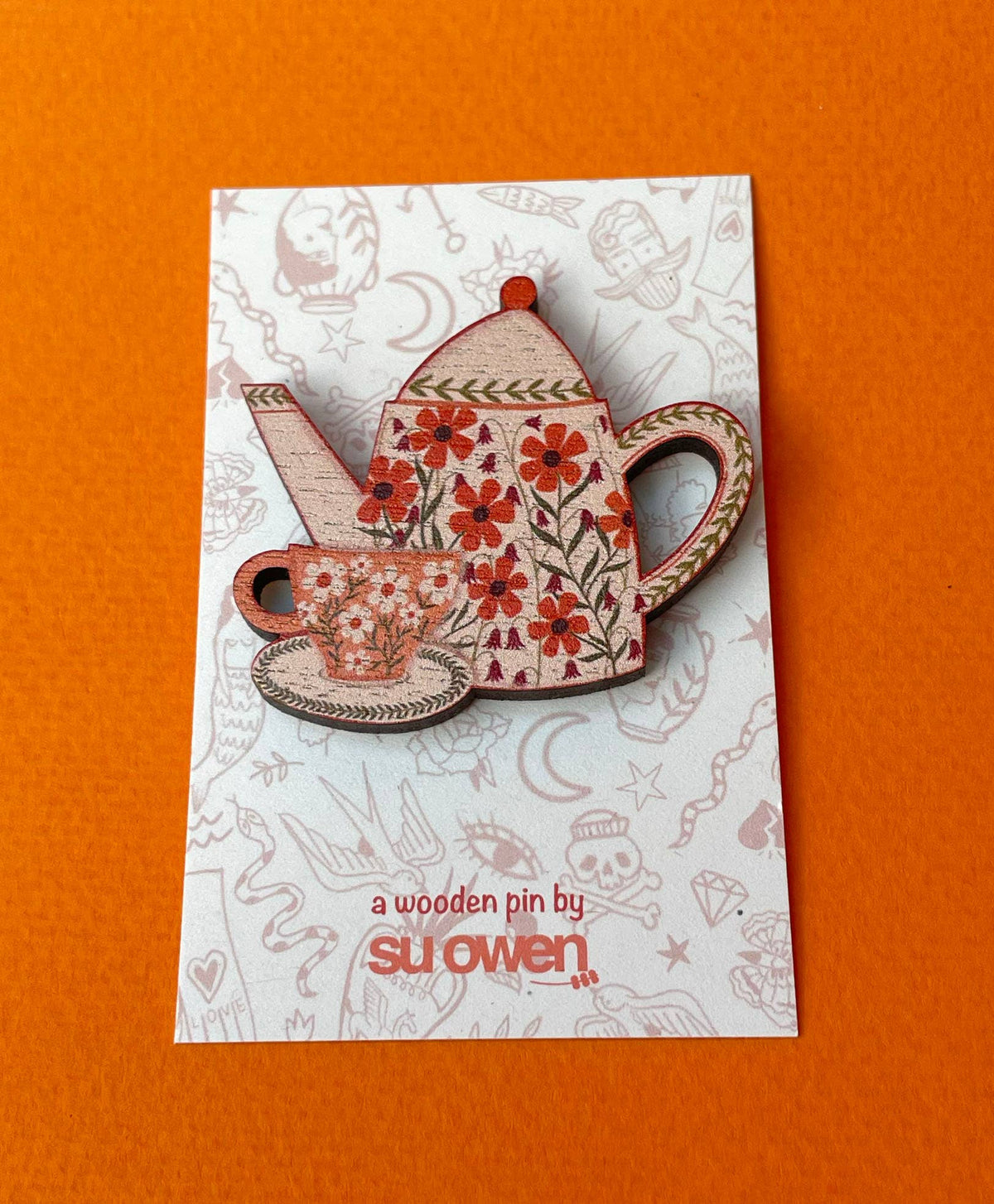A wooden tea set pin featuring a teapot and teacup with red floral patterns, displayed on a branded backing card against a solid orange background.