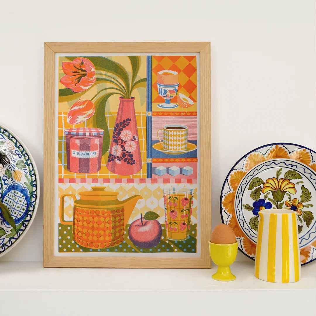Framed colorful artwork of a kitchen scene with plates and a teapot on a white wall.