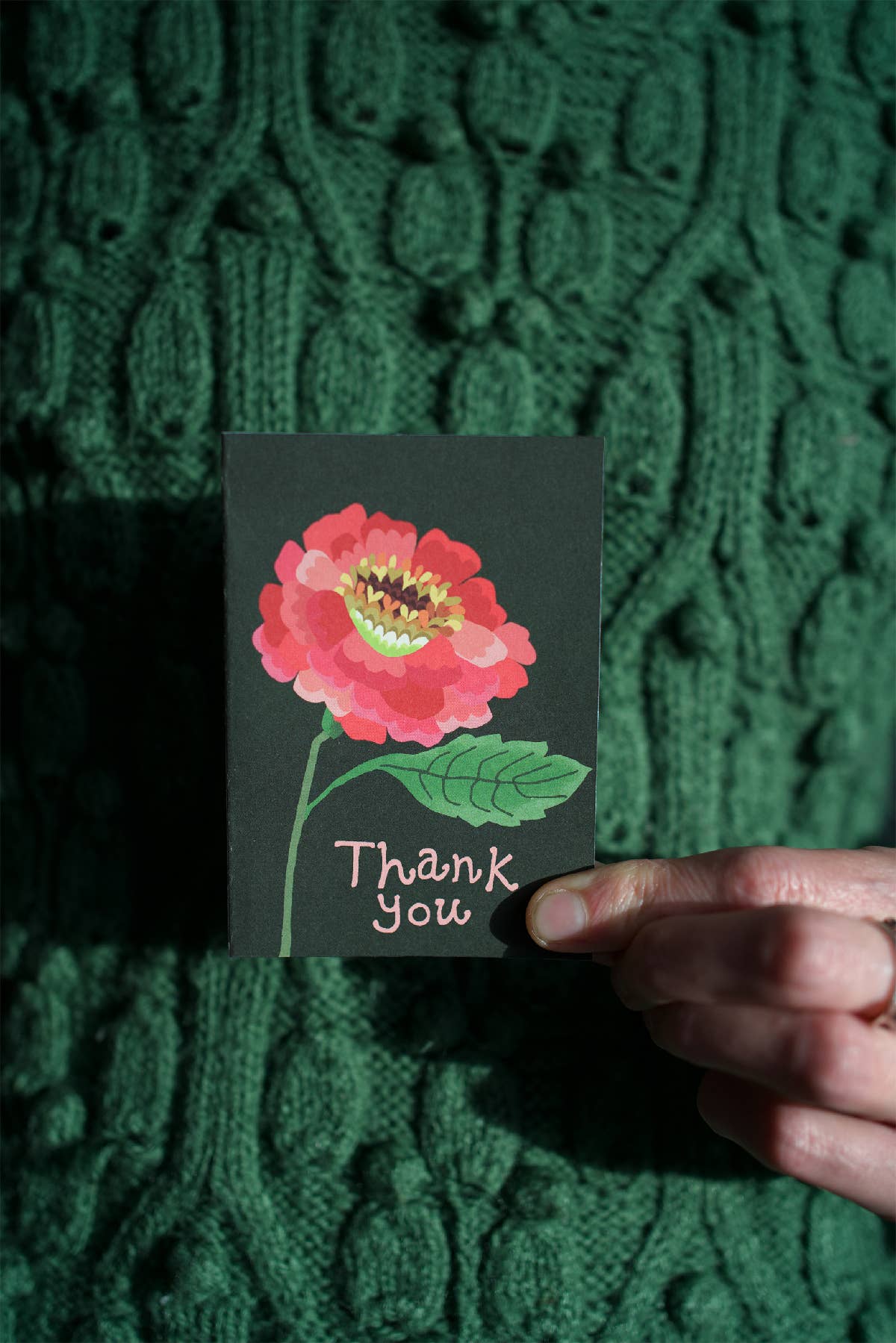 Card with a pink flower and 'Thank You' text held against a green textured background.