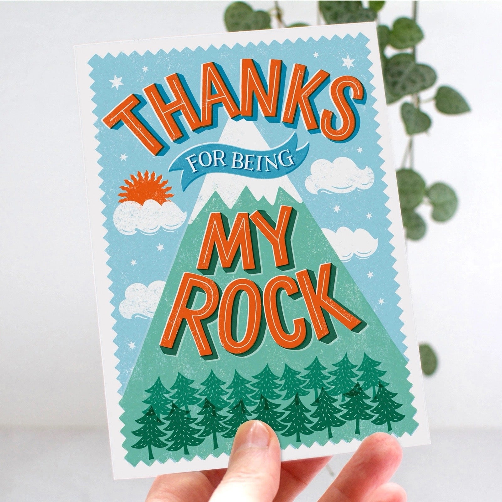 Card with mountain design and text 'Thanks for being my rock' held by a hand.