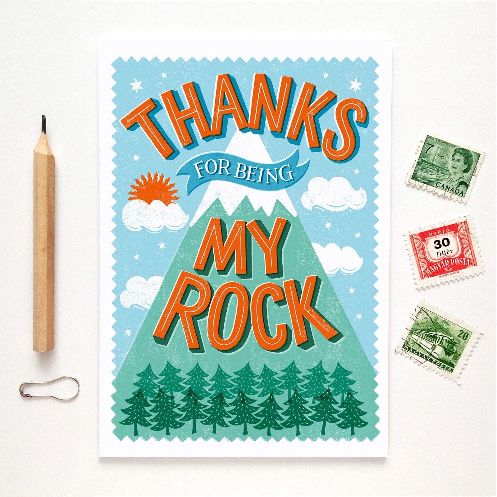 Card with mountain design and text 'Thanks for being my rock' on a white background with a pencil and stamps.