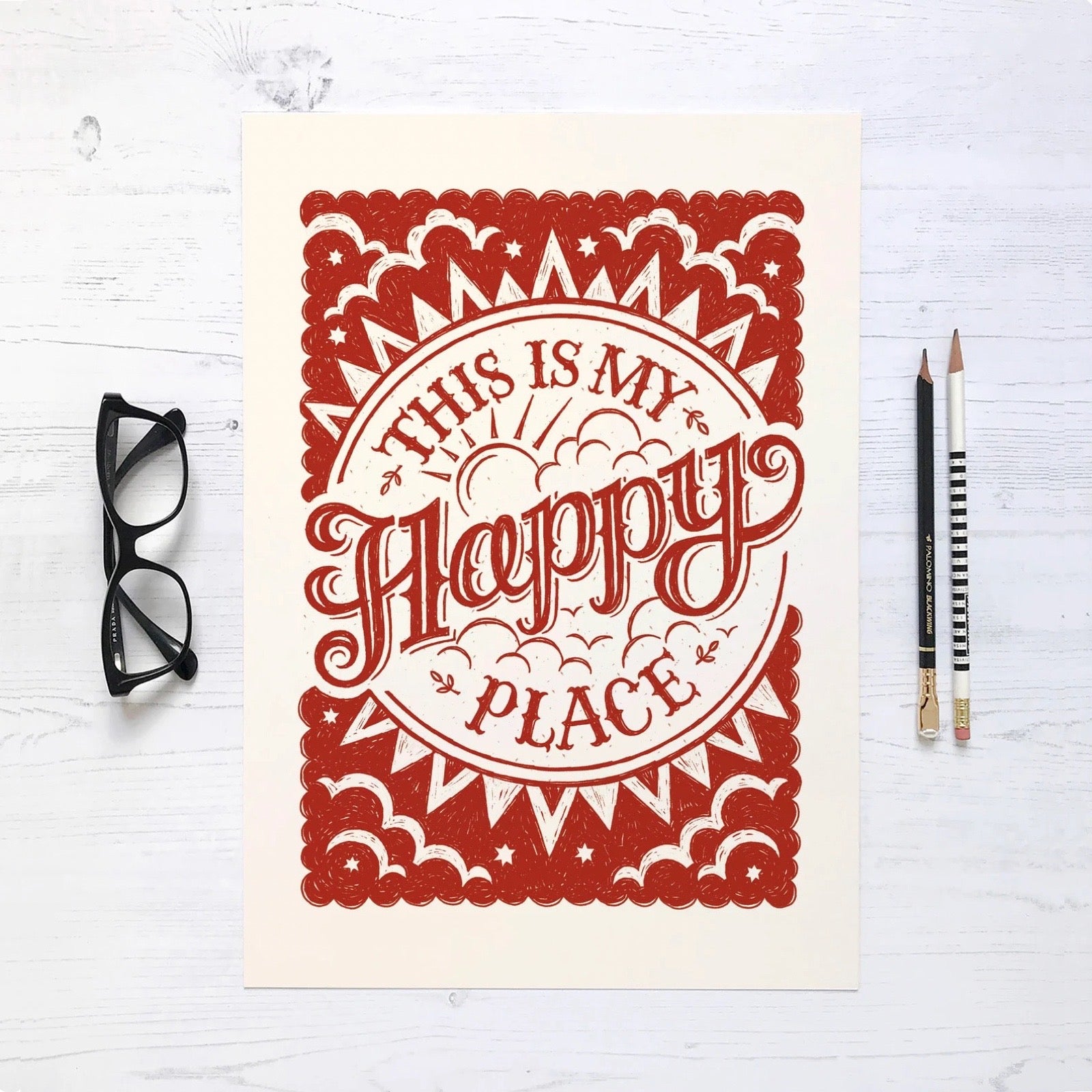 Decorative print with 'This is my happy place' text on a white surface with glasses and pencils.