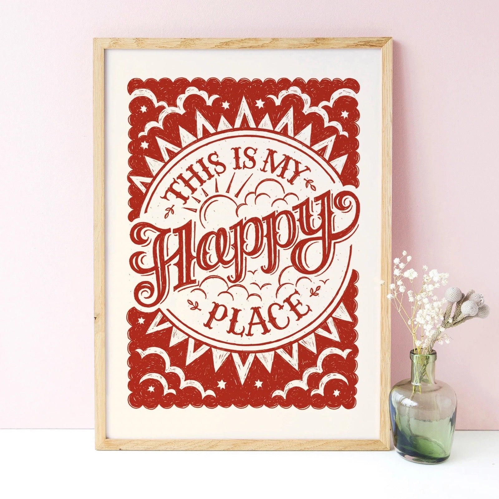 Framed red and white decorative print with 'This is my happy place' text on a pink wall, next to a flower in a vase.