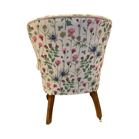 A cream linen armchair with a thistle floral pattern in pinks and blues, featuring green foliage. The chair has a button-back design, cream binding, and is set on mahogany legs with gold castors.