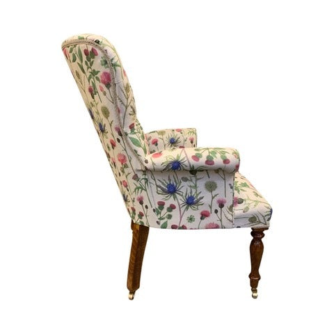 A cream linen armchair with a thistle floral pattern in pinks and blues, featuring green foliage. The chair has a button-back design, cream binding, and is set on mahogany legs with gold castors.