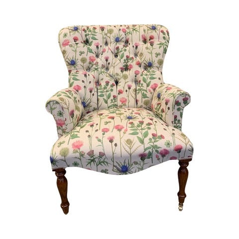 A cream linen armchair with a thistle floral pattern in pinks and blues, featuring green foliage. The chair has a button-back design, cream binding, and is set on mahogany legs with gold castors.