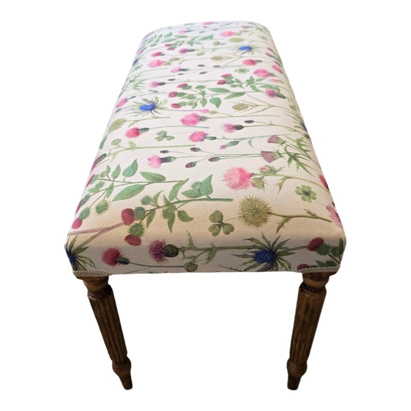A rectangular bench with cream linen upholstery, featuring a floral pattern with pink and purple thistles and green foliage, and mahogany legs.