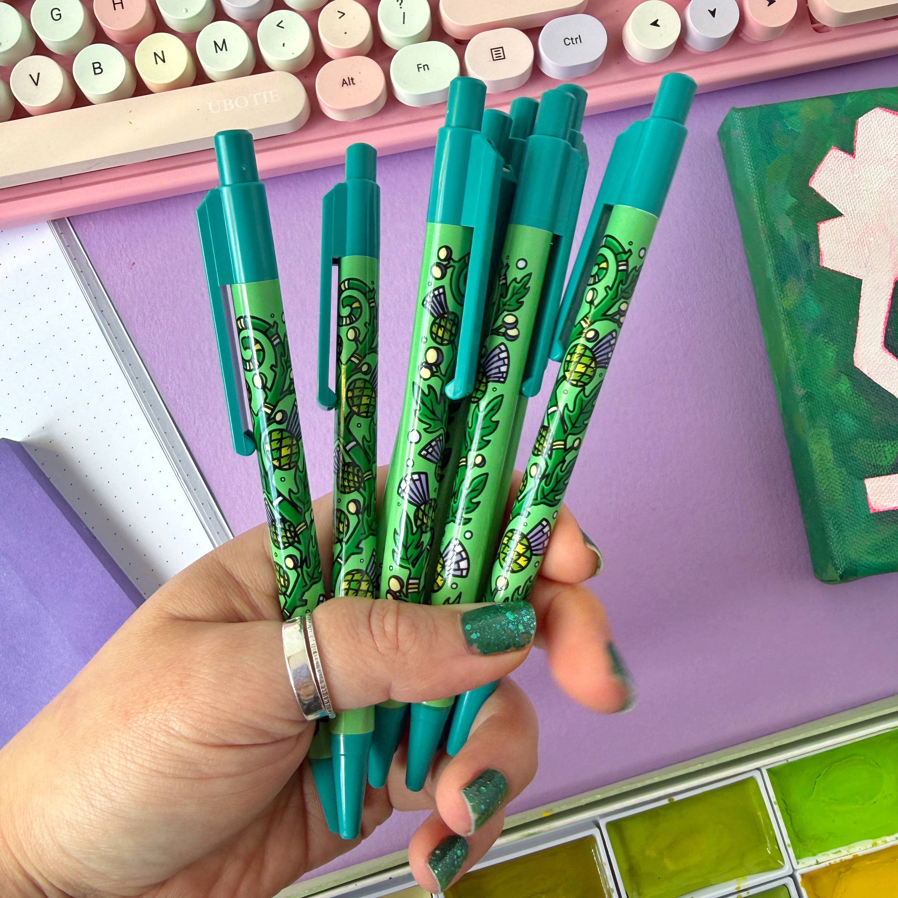 Hand holding green pens with a colourful keyboard and stationery items in the background.