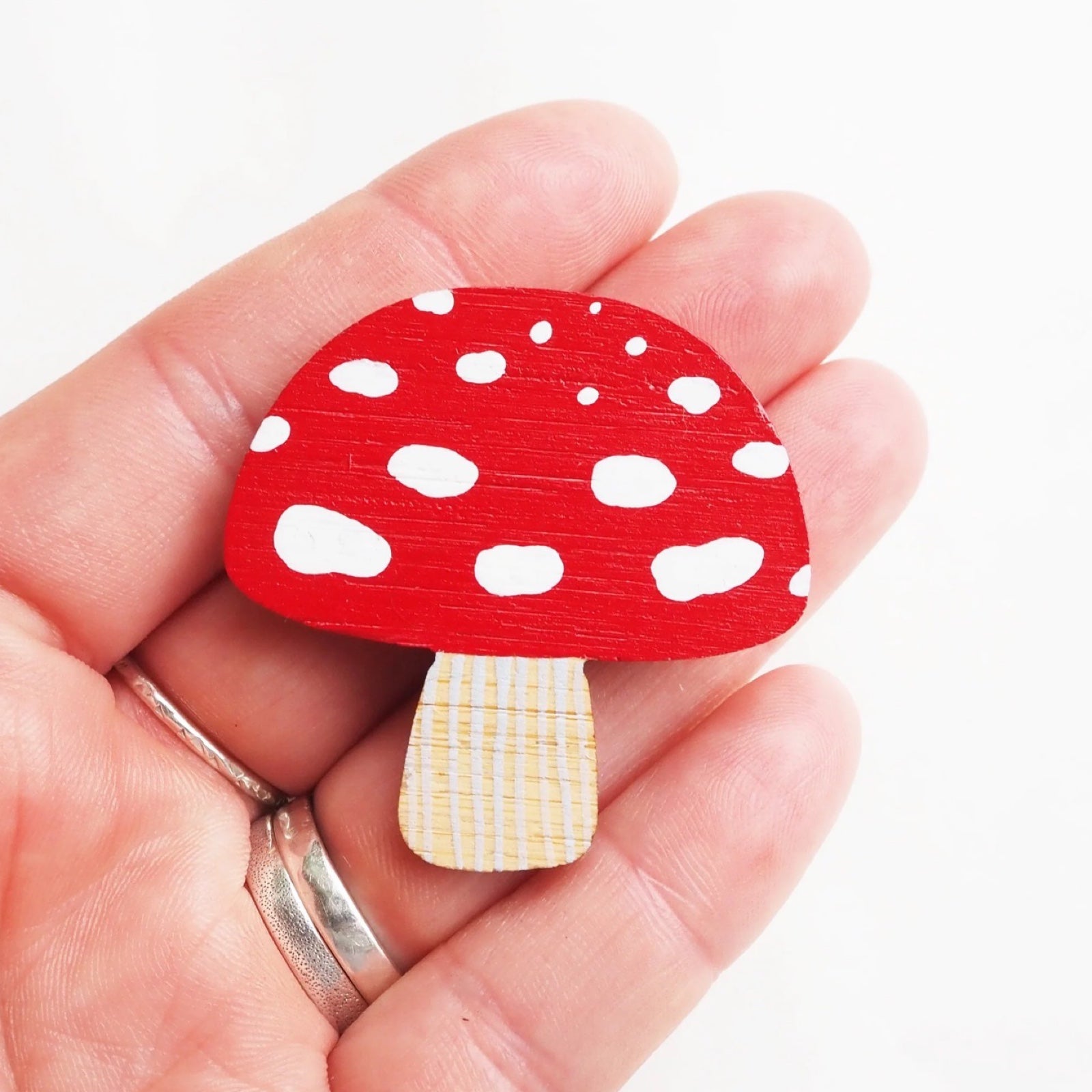 Toadstool-shaped brooch is made from lightweight and hardwearing bamboo, hand-painted in red and white and sealed with a water-based satin varnish, being held on a hand.
