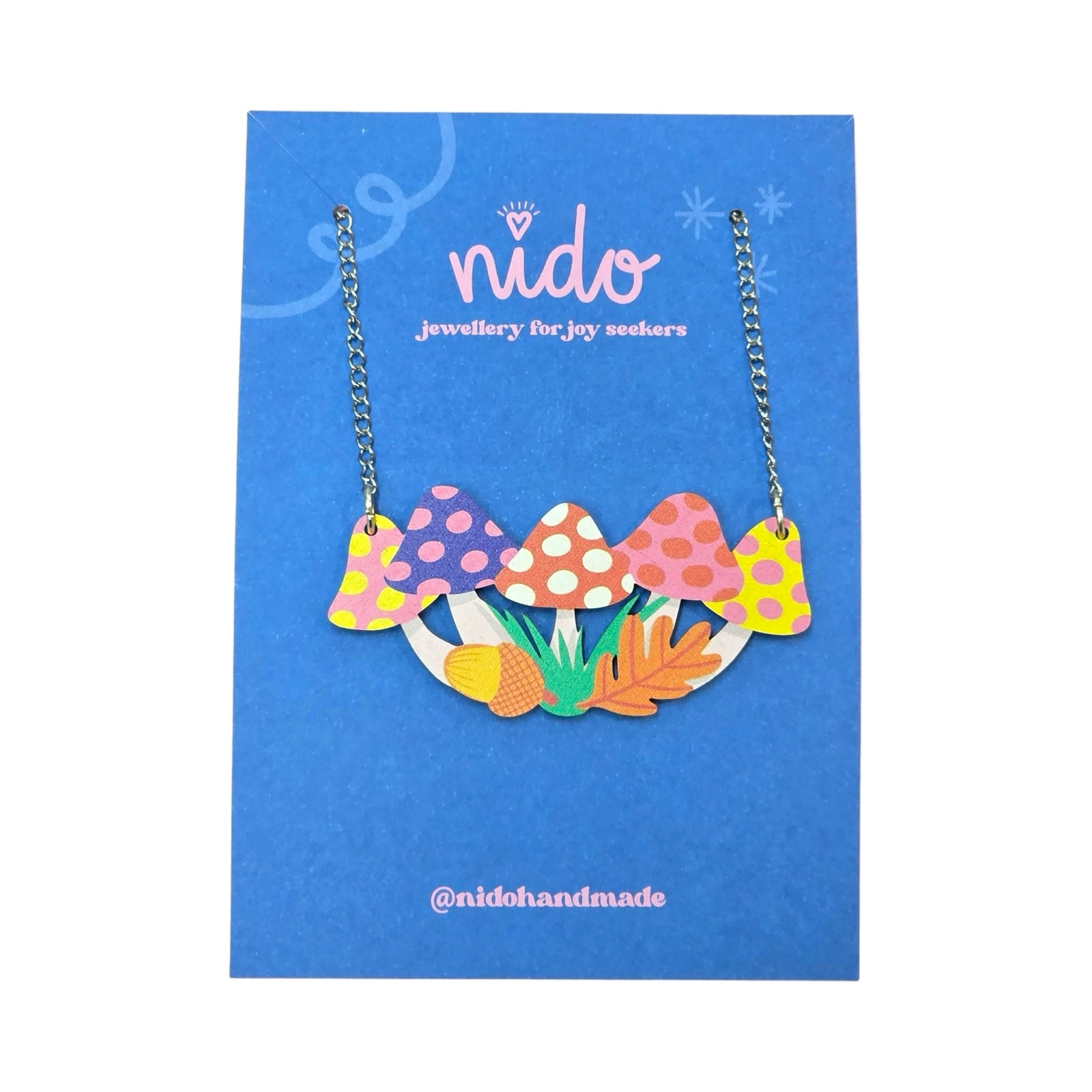 This wooden toadstool necklace features yellow, blue and pink toadstools with an acorn, grass and a leaf in the centre of their stalks, presented on a branded blue backing card.