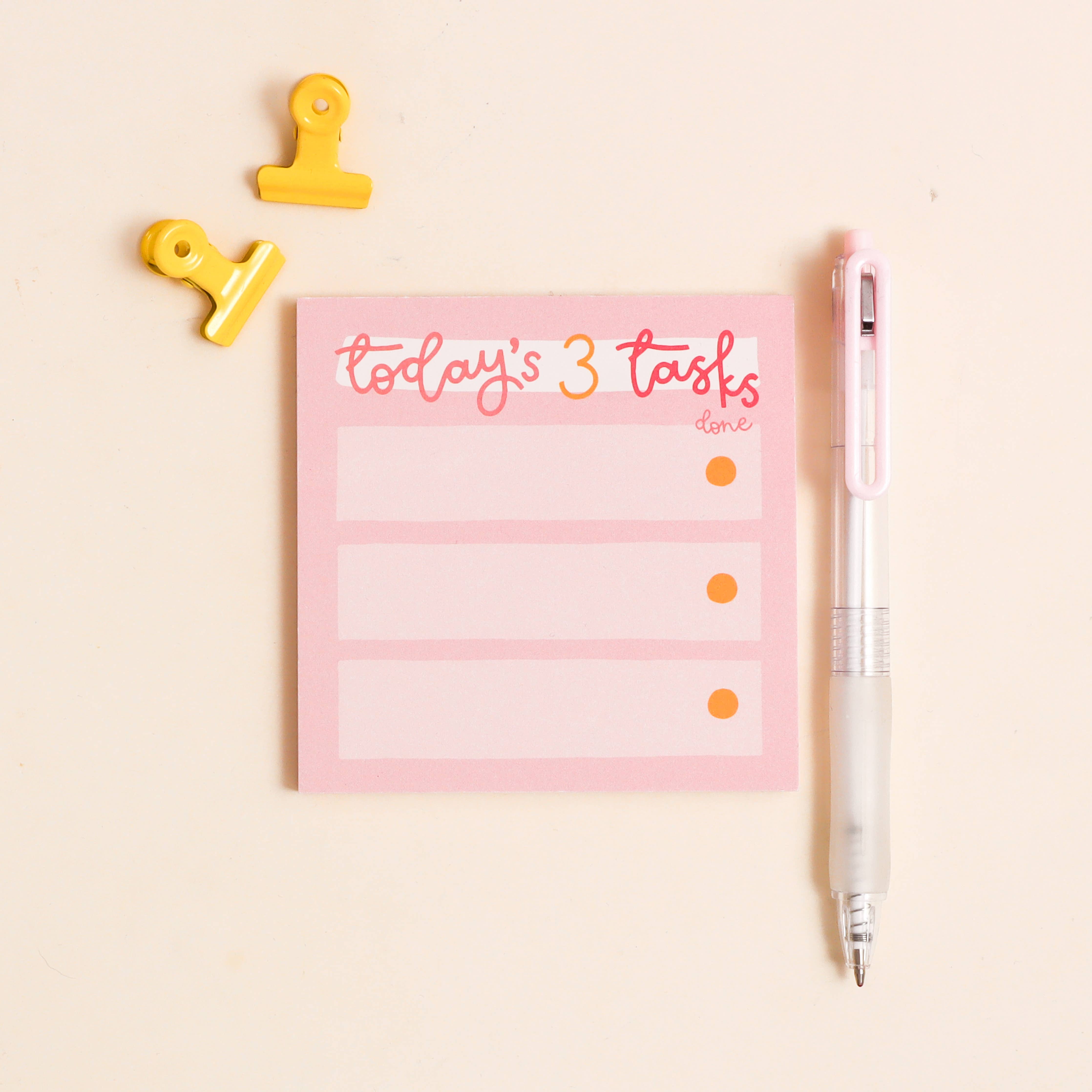 Pink notepad with 'today's 3 tasks' text, yellow clips, and a pen on a light beige background.