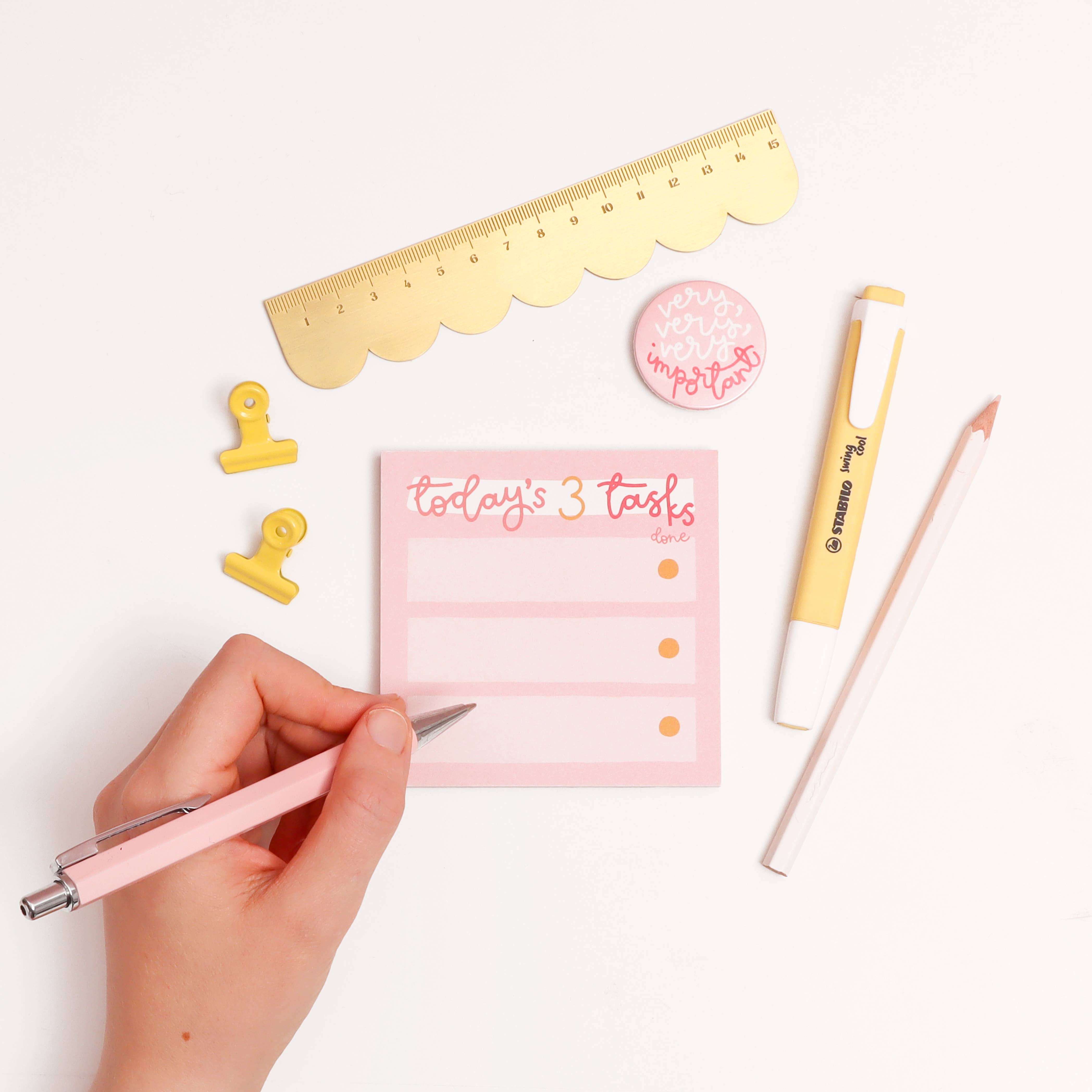 Hand holding a pen near a pink notepad with 'Today's 3 Tasks' on a white background with stationery items.