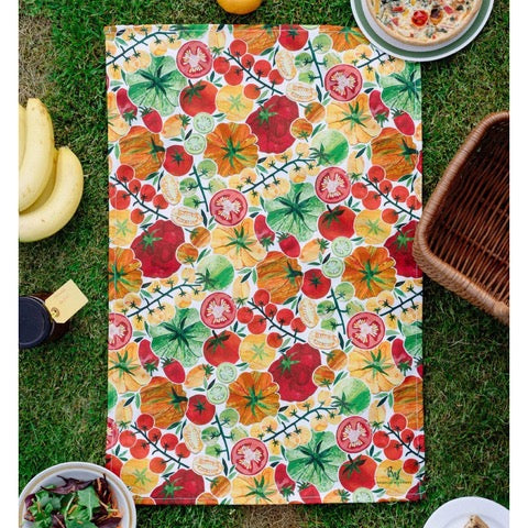 Tea towel featuring red, orange and yellow tomatoes with green foliage on a white background.