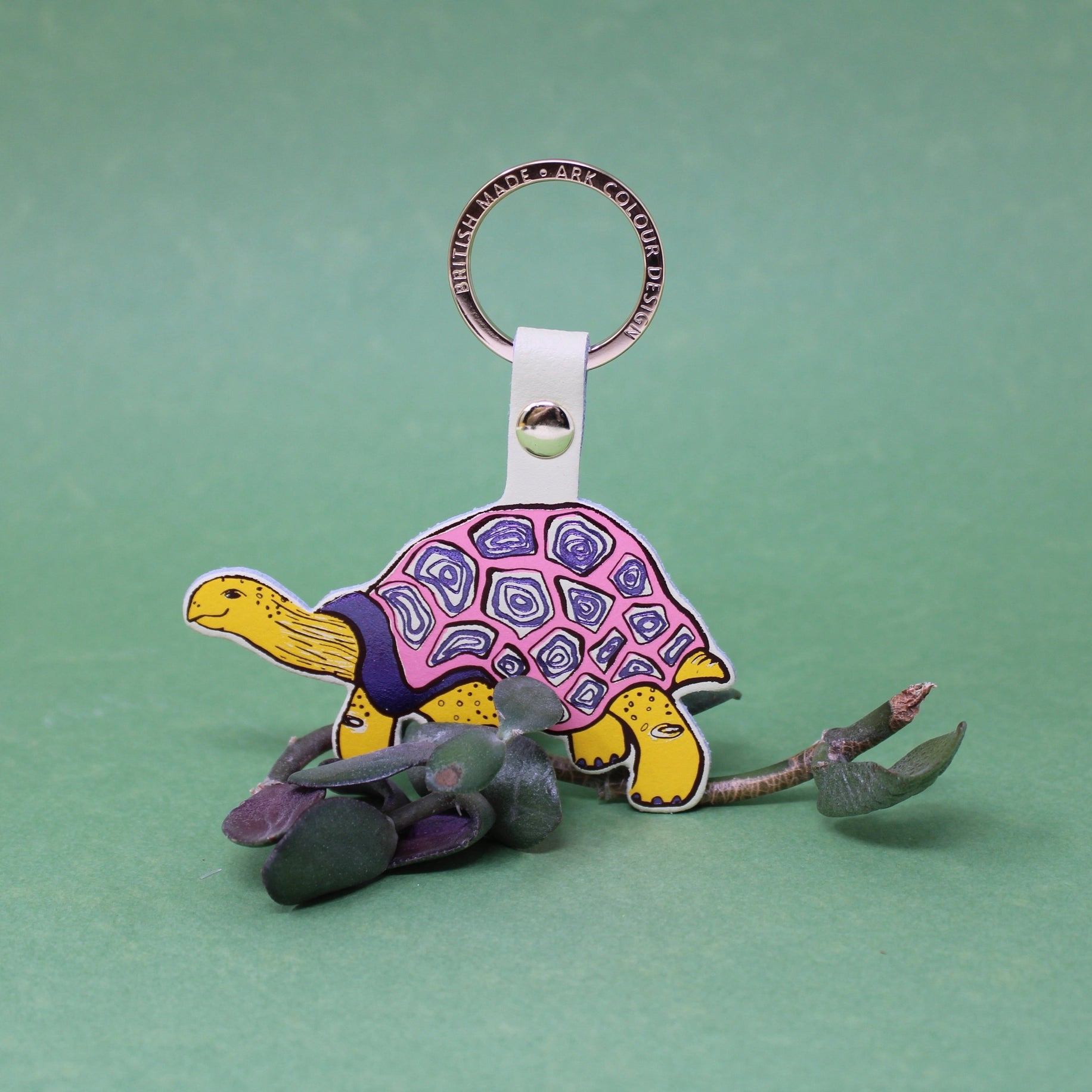Tortoise-shaped keychain with a pink shell  and yellow limbs on a green background, with a branch of leaves.
