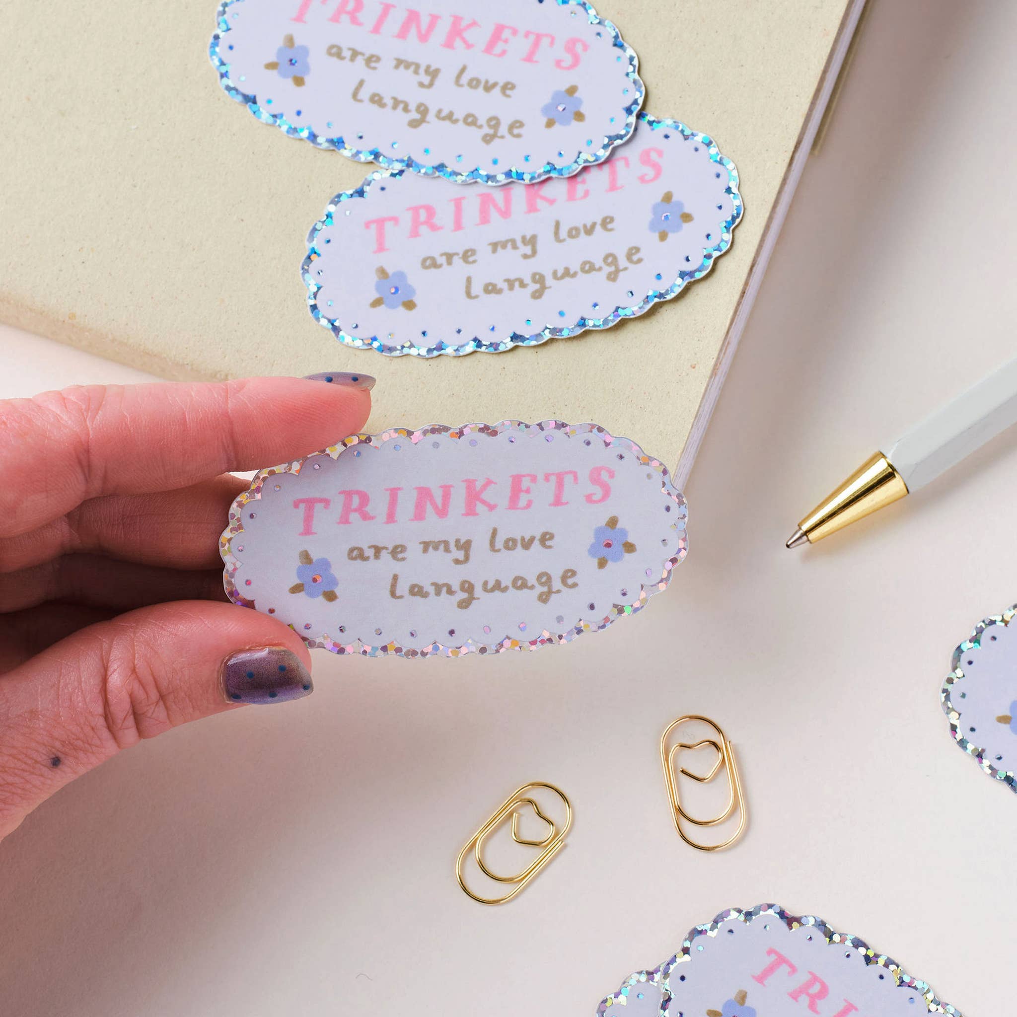 Hand holding a decorative sticker with 'TRINKETS are my love language' text, surrounded by more stickers and a pen on a light surface.