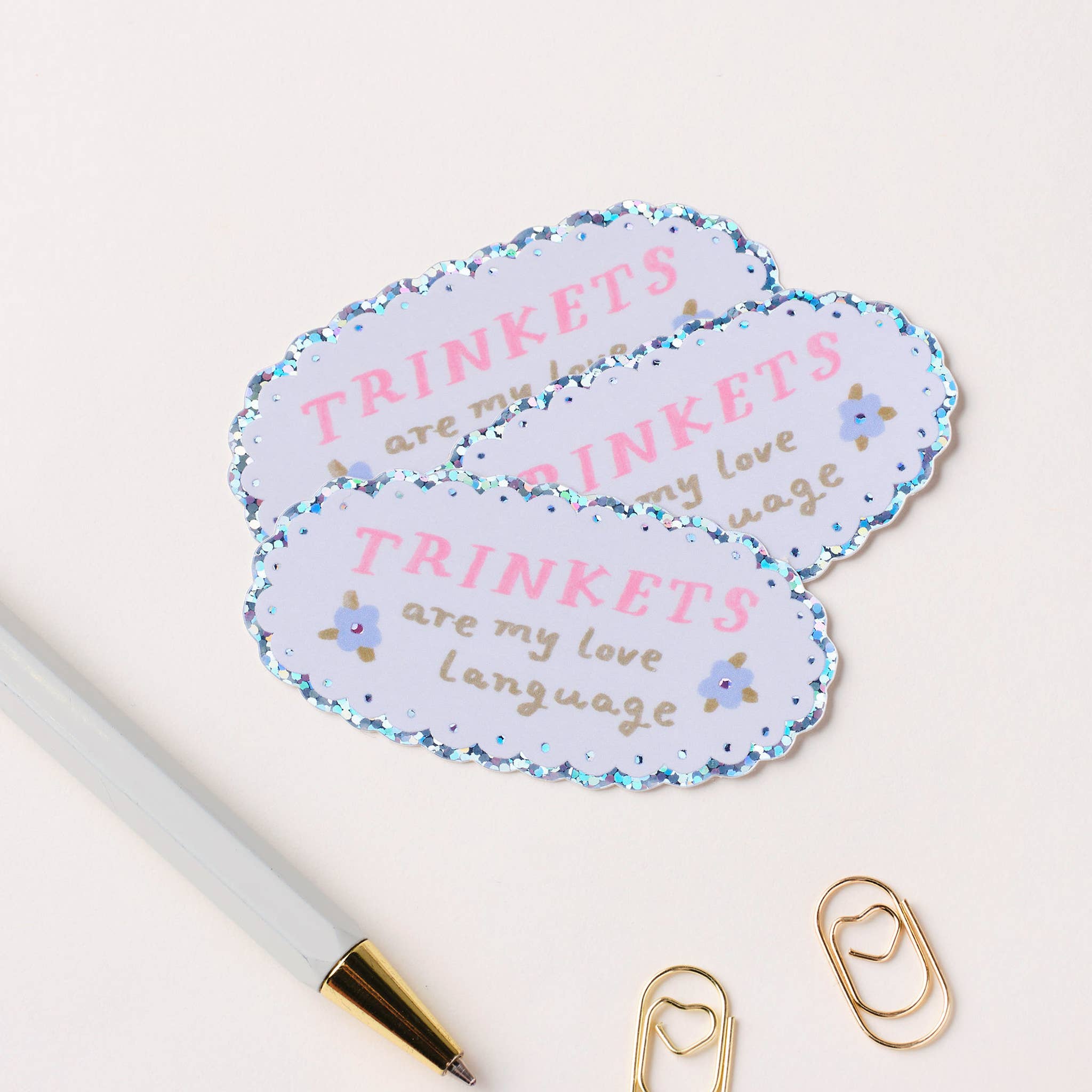 Decorative stickers with 'TRINKETS are my love language' text on a white background with a pen and paper clips.