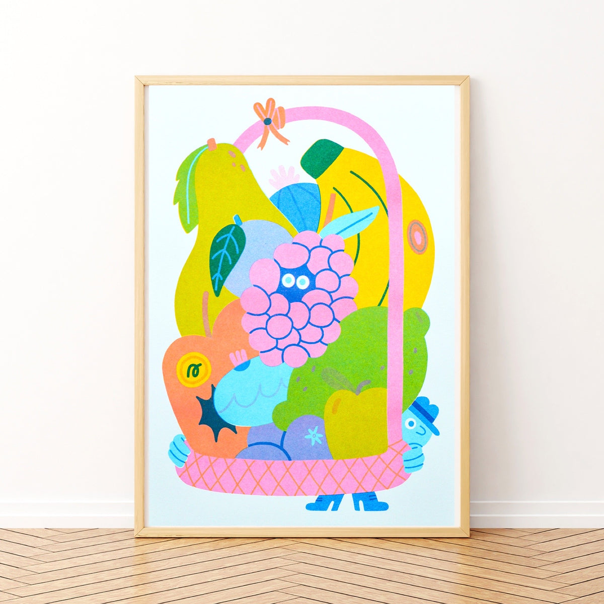 A framed art print featuring a whimsical, colourful illustration of a fruit basket with fruits and a blue character, leaning against a white wall on a light wood floor.