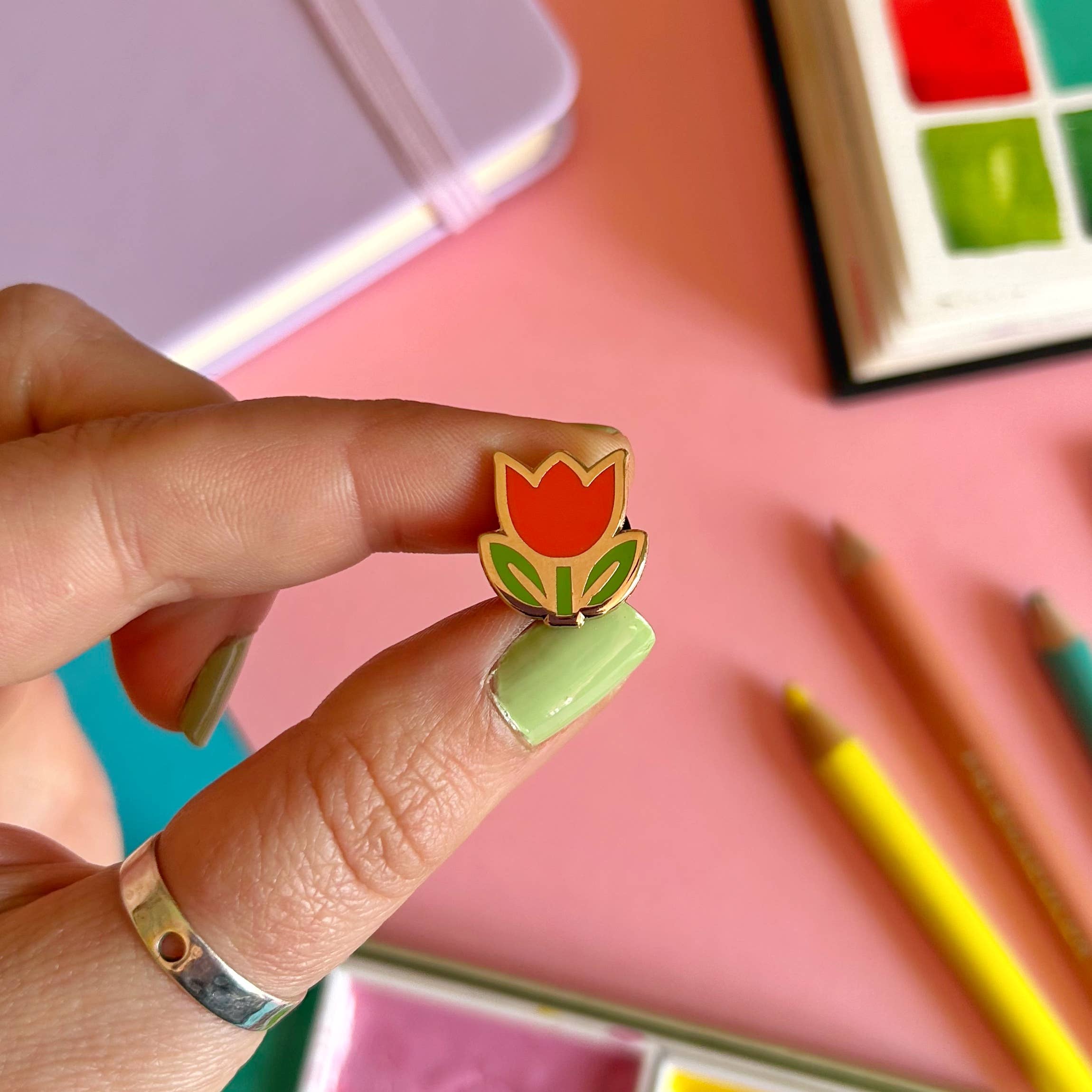 Hand holding a tulip-shaped pin with watercolour paints and pencils in the background.