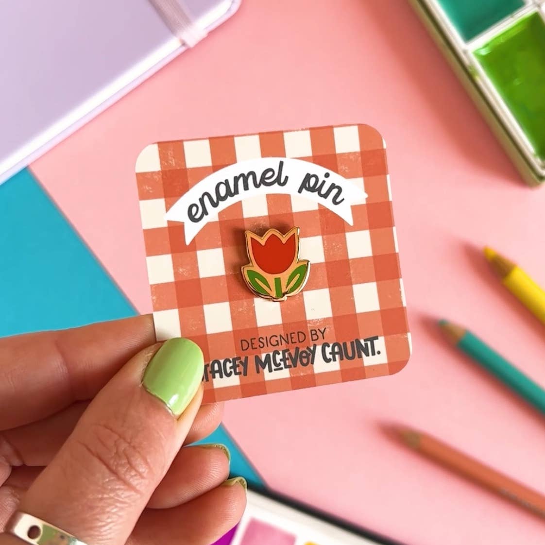Enamel pin of a tulip on a checkered card held by a hand with green nail polish, against a colourful background.