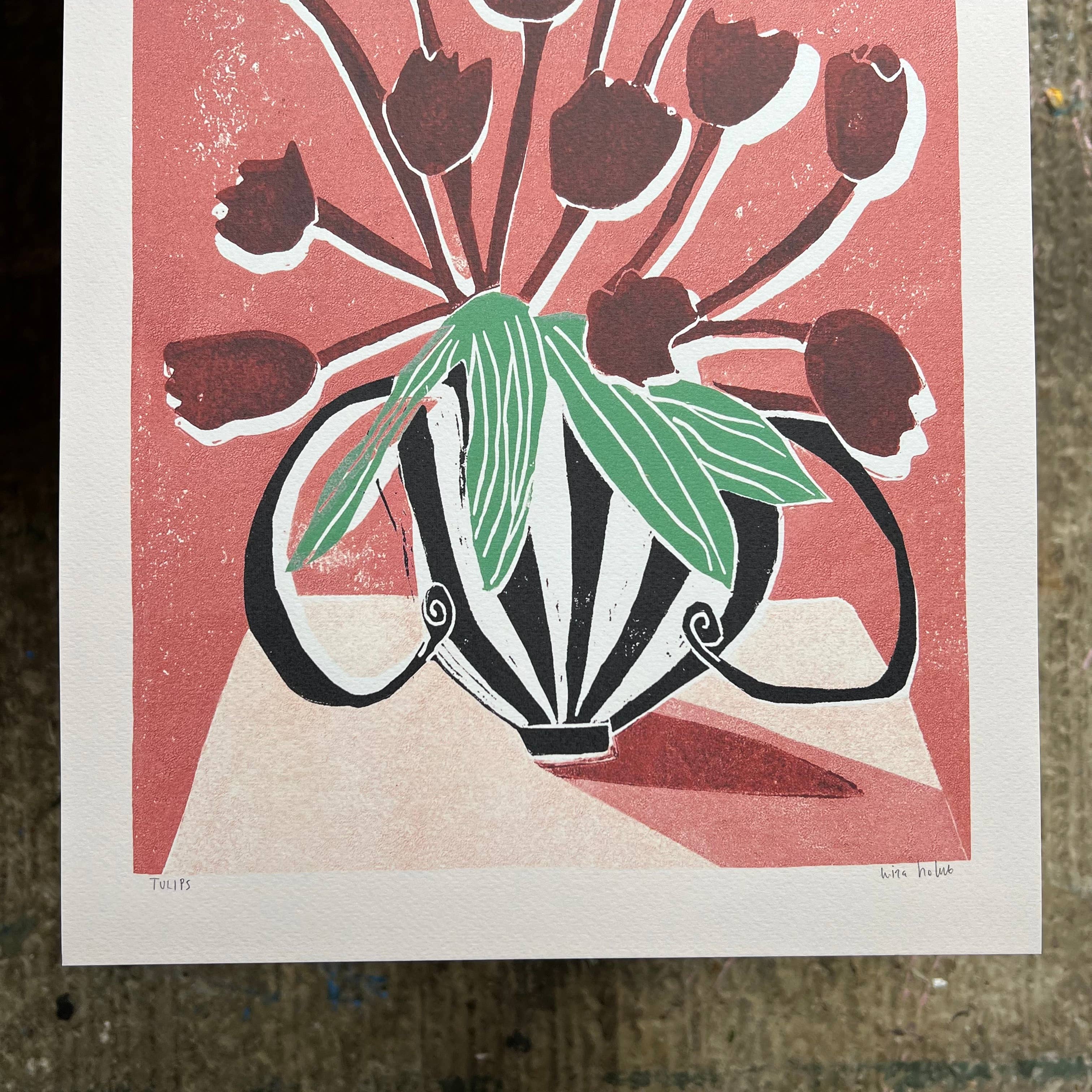 An art print featuring a stylised floral illustration with dark red tulips, green leaves, and a black-and-white striped vase on a coral background, photographed from above on a textured surface.