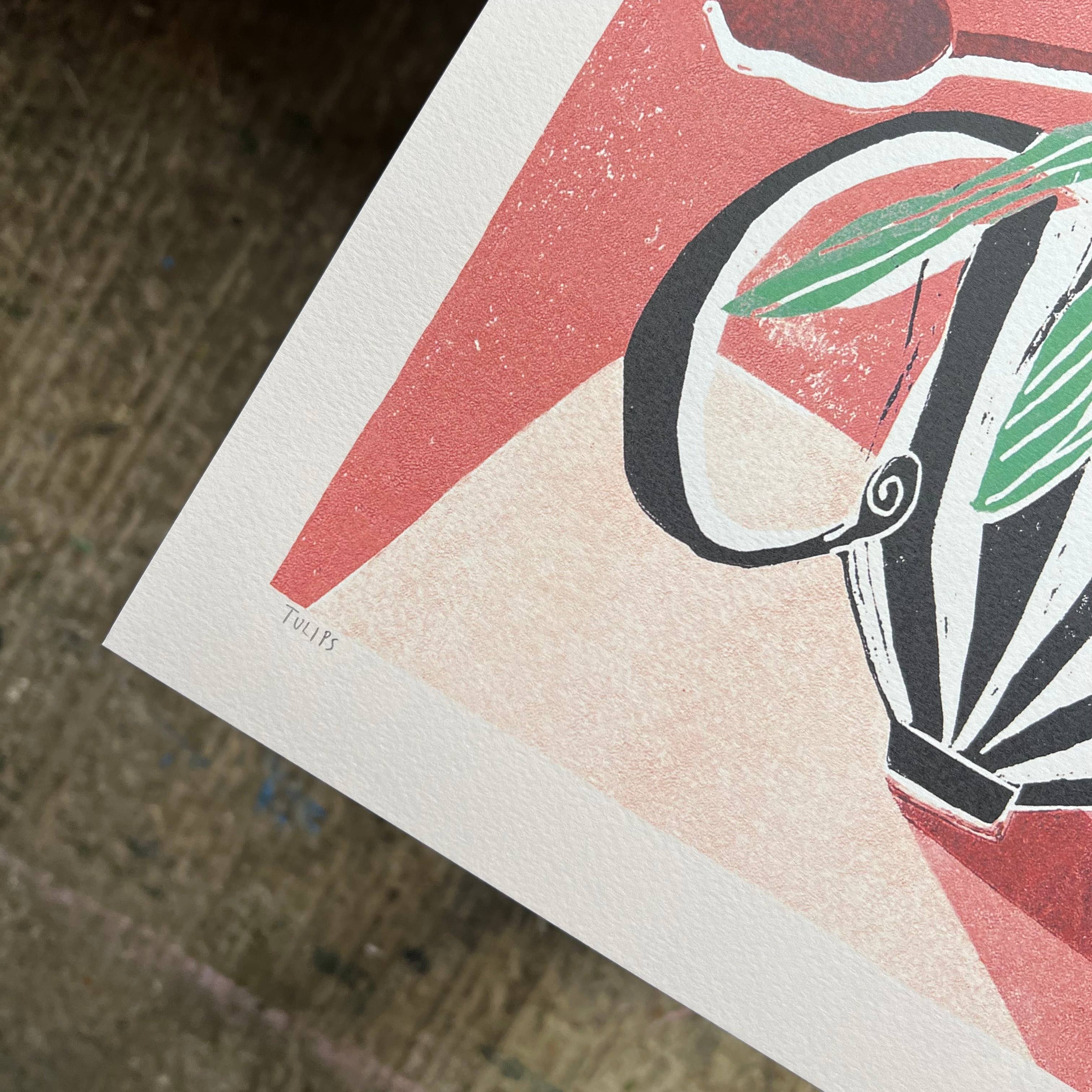 A close-up of a printed art piece showing a stylised illustration in black, cream, coral red, and green, photographed on a textured brown surface.