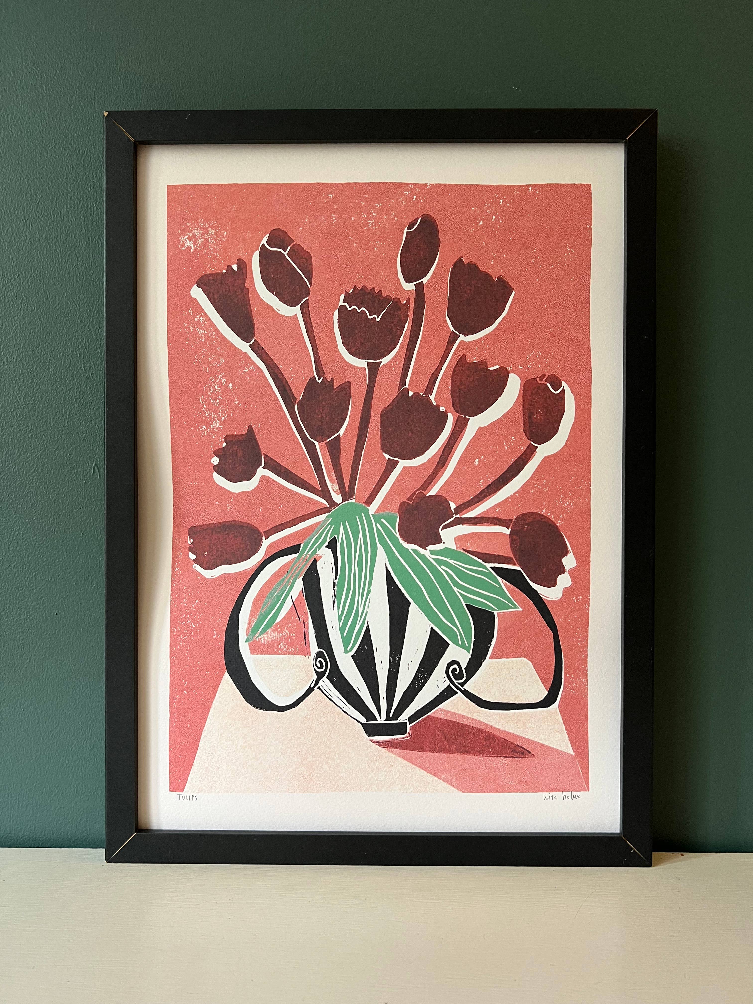 A framed art print featuring a stylised bouquet of dark red tulips with green leaves on a coral background, set in a black frame against a dark green wall.