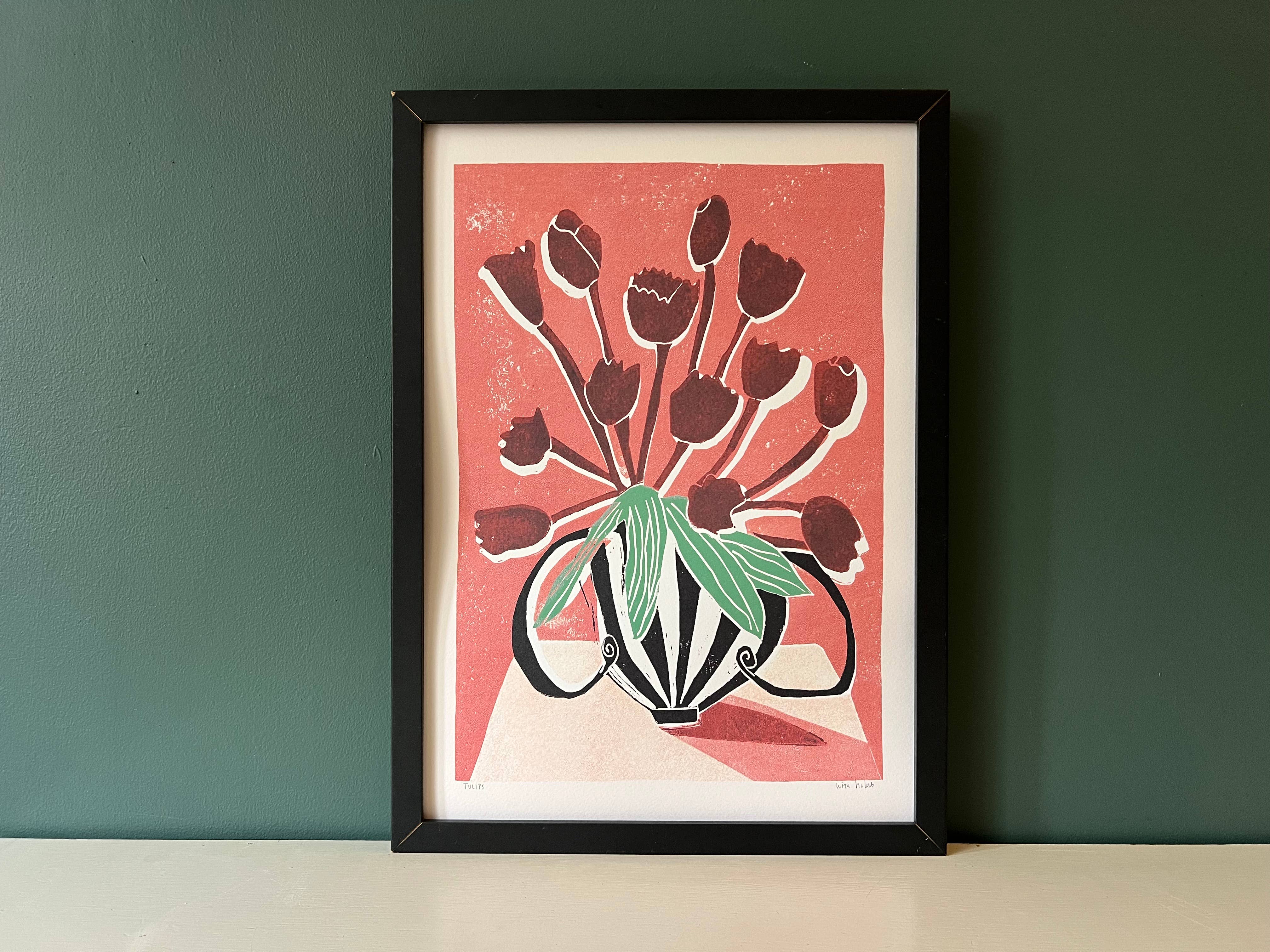A framed art print featuring a stylized bouquet of dark red tulips with green leaves on a coral background, displayed against a dark green wall on a light surface.