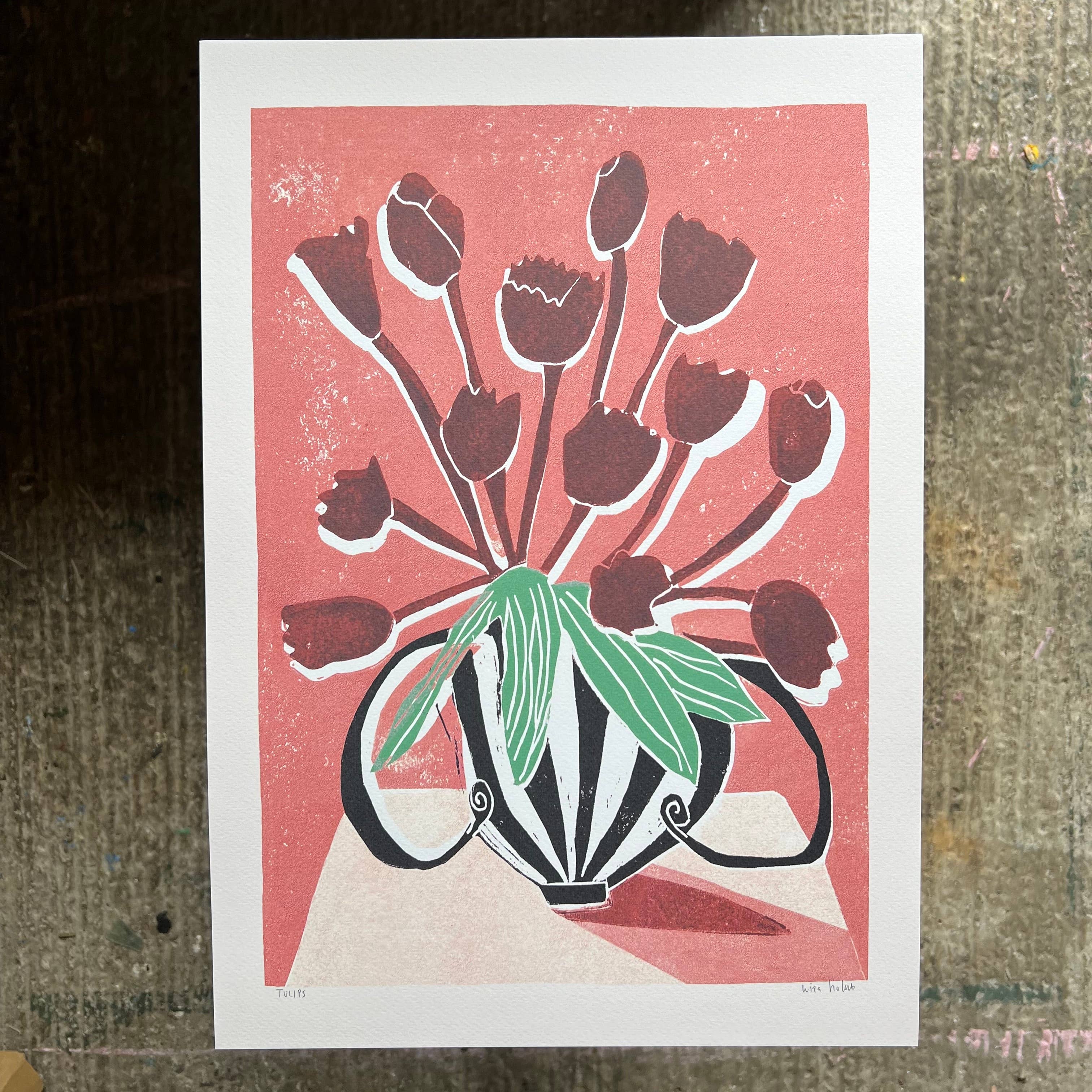 A square art print featuring a stylized floral arrangement of dark red tulips with green leaves in a black-and-white patterned vase, set against a coral pink background. The print has a wide white border and is photographed flat on a textured gray surface.