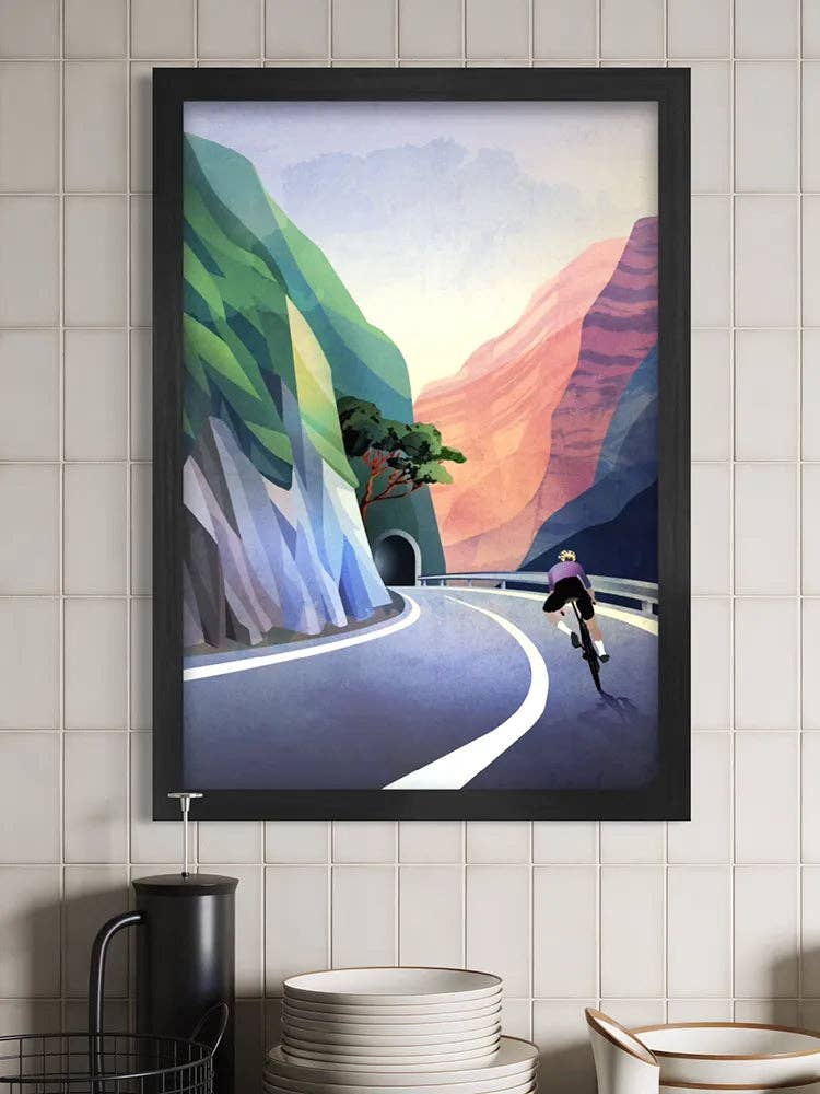 A framed art print depicting a stylised cyclist on a winding mountain road, hanging on a white tiled wall above kitchen items.
