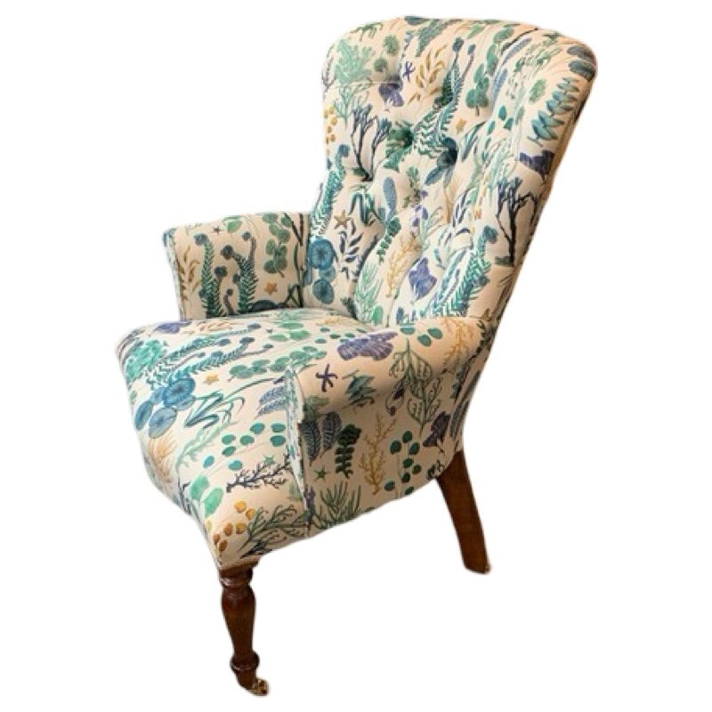 A cream linen fabric upholstered armchair with a floral pattern that includes seaweeds and watery plants in shades of blue and green, featuring a button-back design, mahogany stained legs, and gold castors.