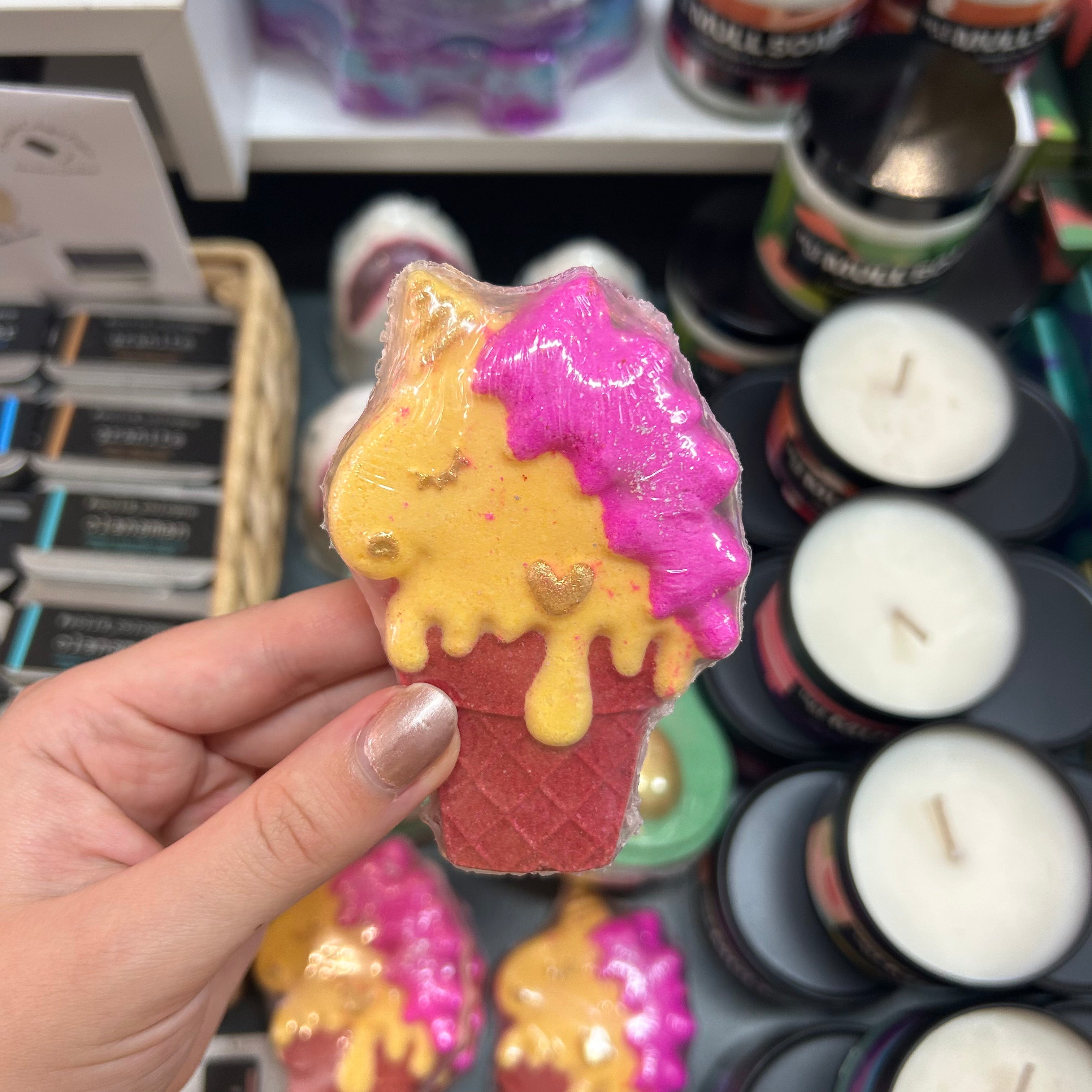 A hand holding a novelty ice cream cone-shaped bath bomb or soap, featuring a yellow body with pink dripping frosting and a red waffle-cone base, with candles and other products blurred in the background.