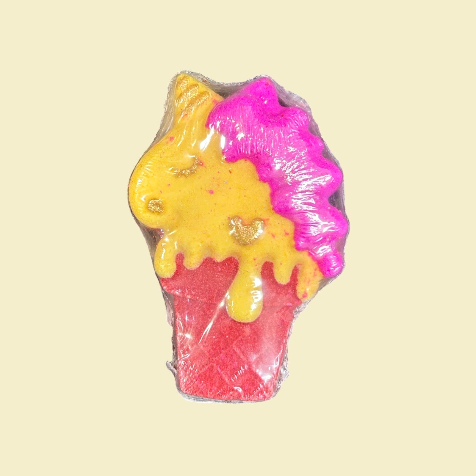 A small bath bomb shaped like a unicorn head into a red cone, wrapped in clear plastic on a pale cream background. The design features a yellow horn and face with pink hair, and glittery red and yellow colours.