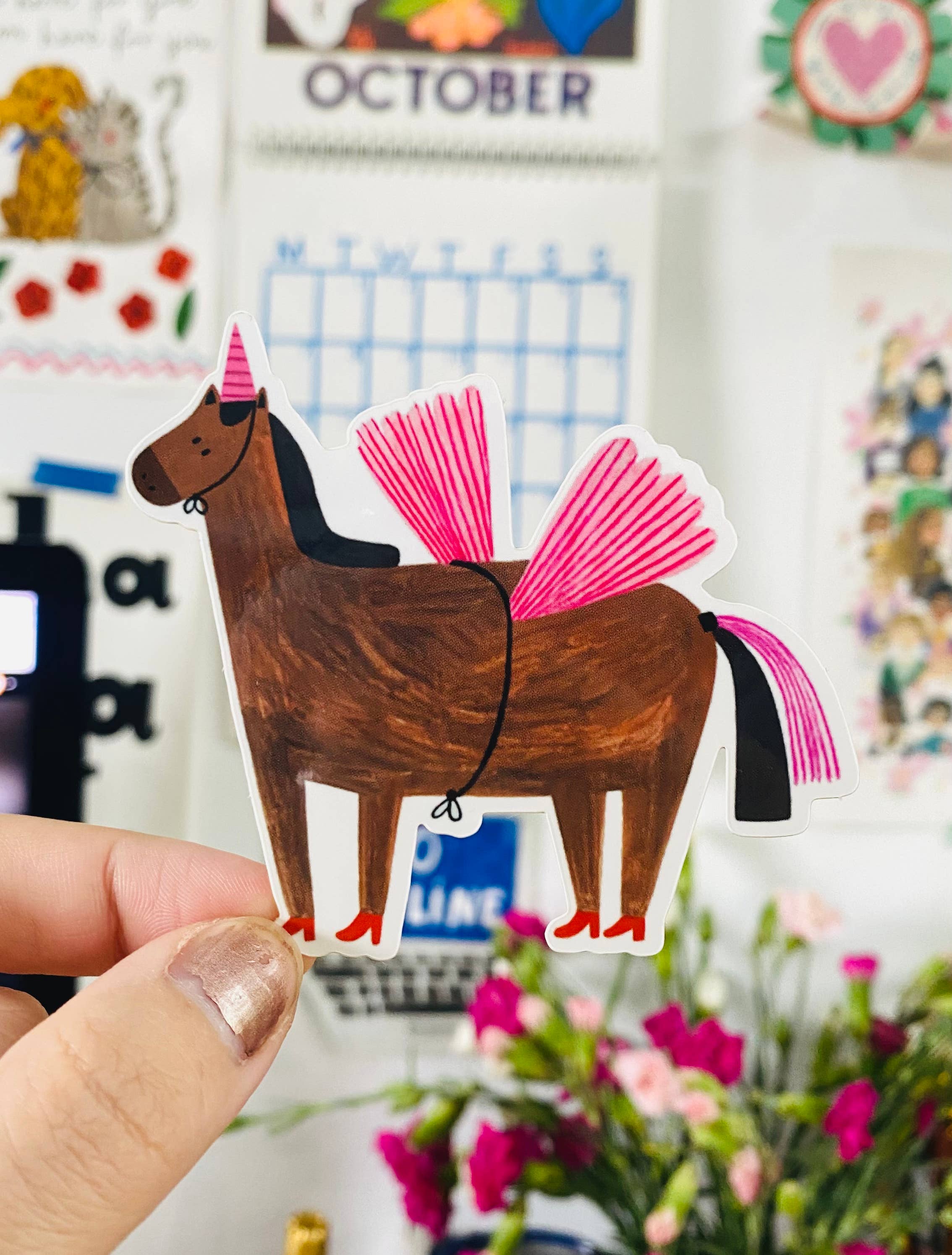 Horse-shaped sticker with a pink tail held by a hand in front of a colorful background.