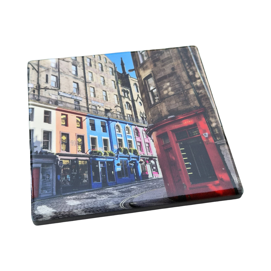 A square coaster featuring a colorful street scene print of Edinburgh, with painted buildings in blue, pink, and red, and a cobblestone street, shown on a plain white background.