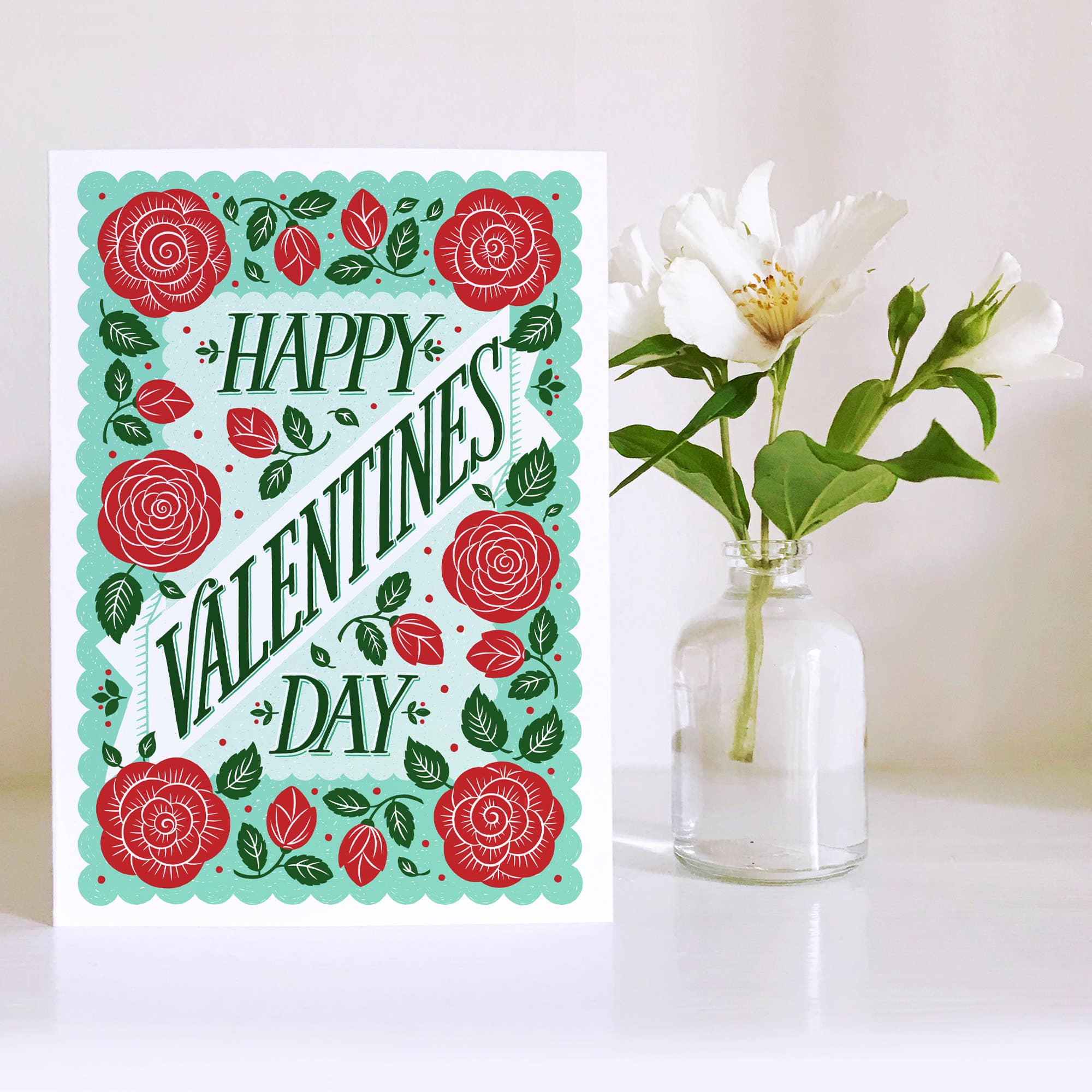 Valentine's Day card with red roses and green leaves next to a vase with white flowers on a white background.