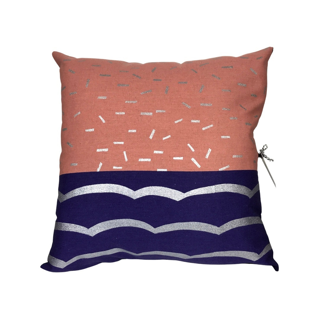 A cushion cover with a geometric design, featuring a purple and silver wave pattern on the bottom half and blush with silver speckles on the top half, and a visible purple zip on the side.