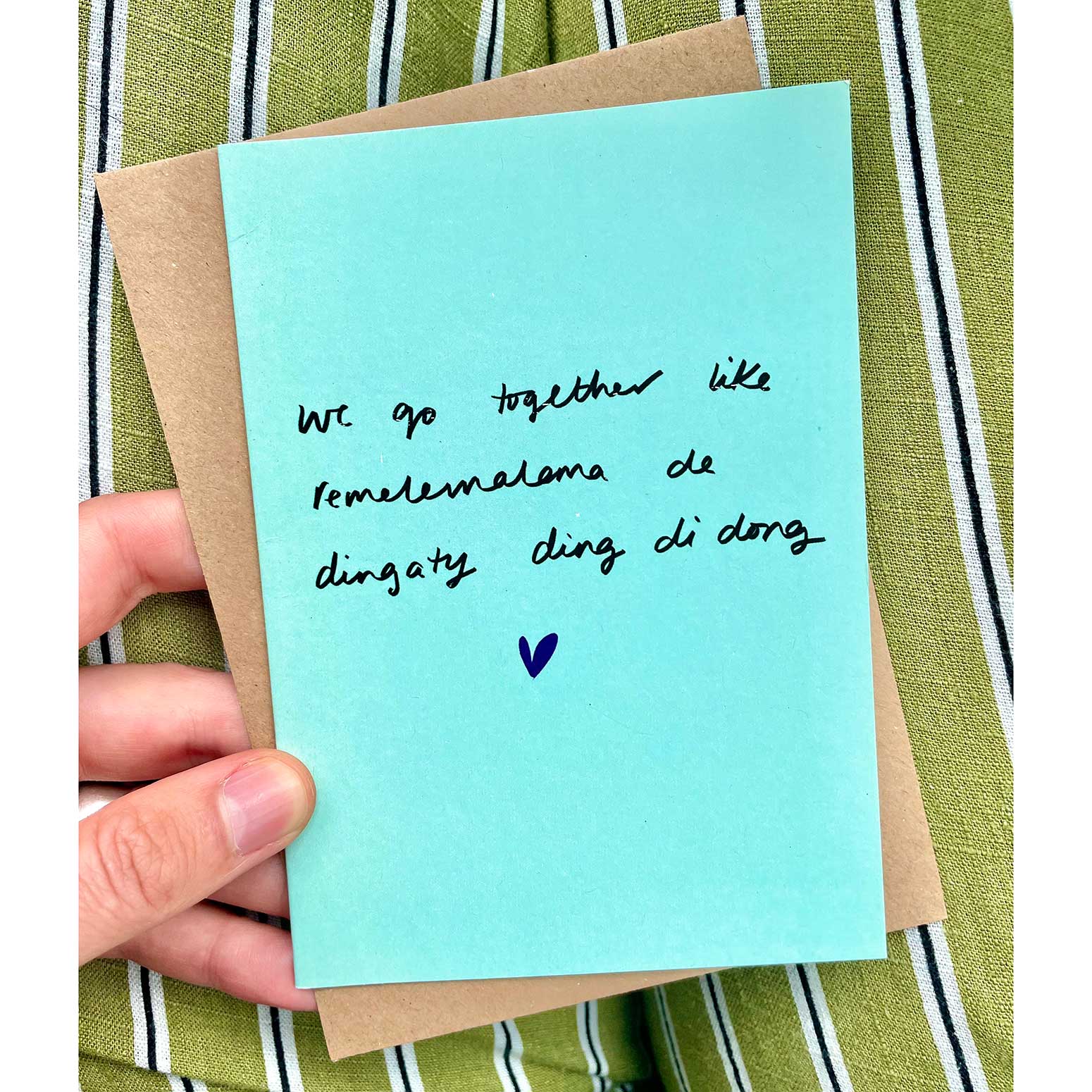 Hand holding a light blue card with text and a heart on a green striped background.