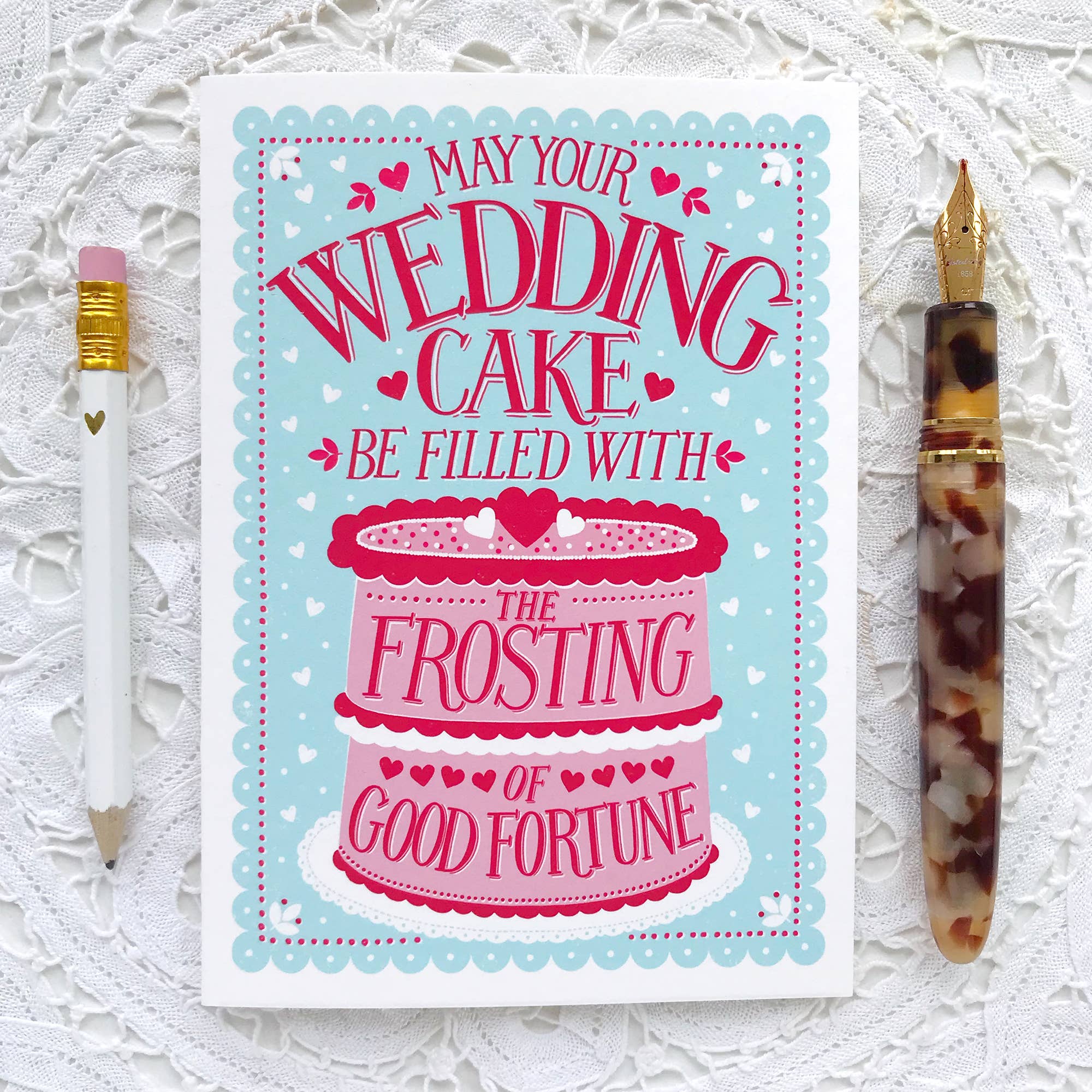 Greeting card with wedding cake design and text, accompanied by a pencil and pen on a textured white background.
