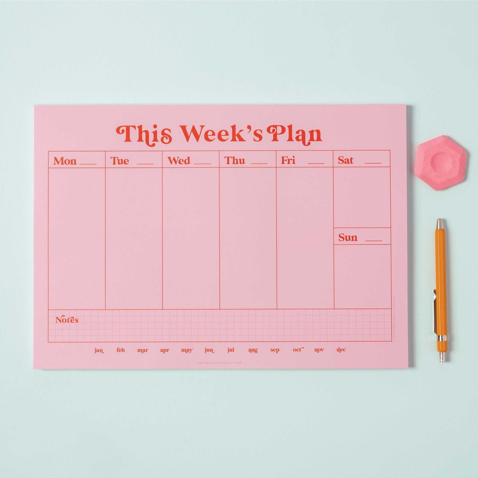 Pink weekly planner with a pen and rubber on a light blue background.