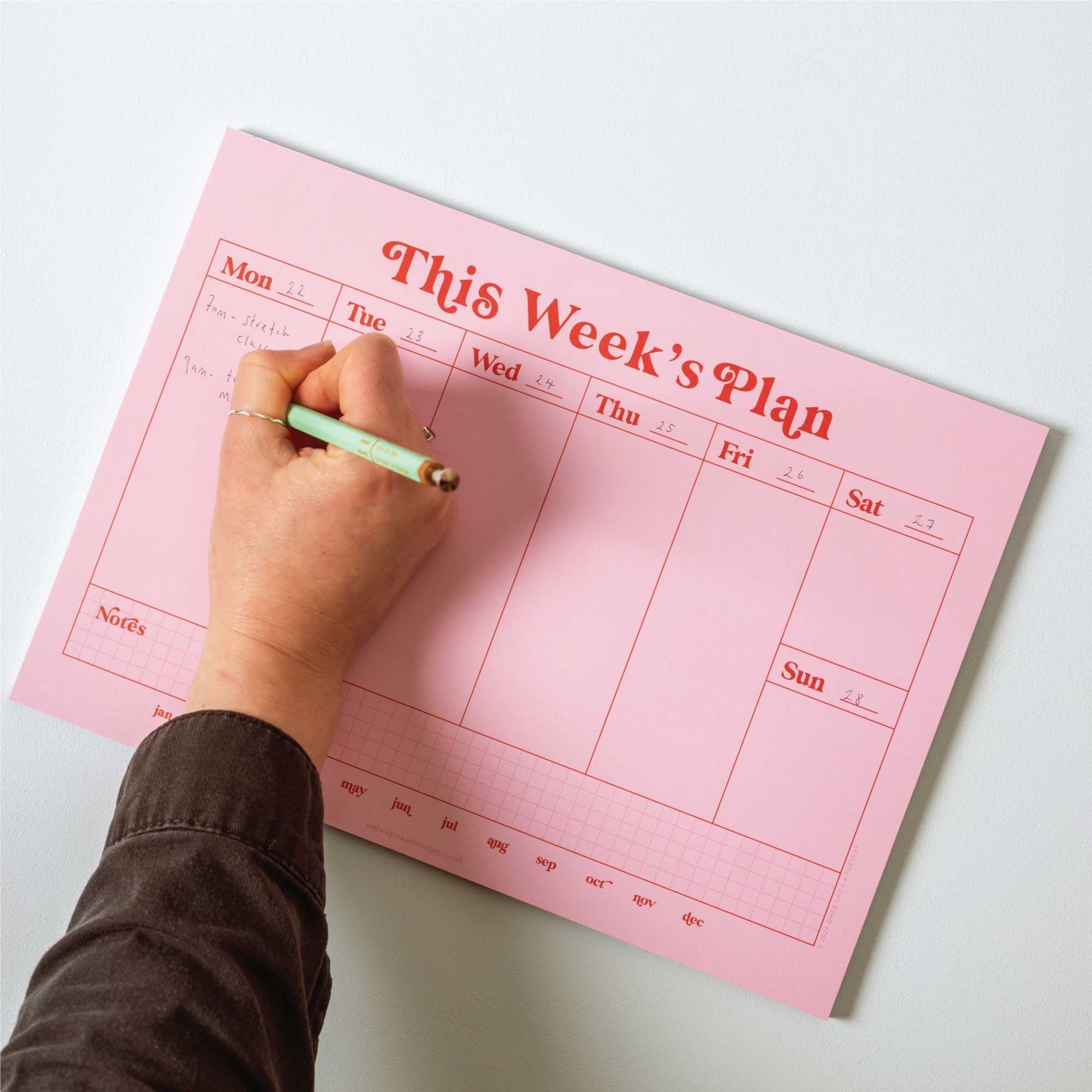 Pink weekly planner with a hand writing on it against a light grey background.
