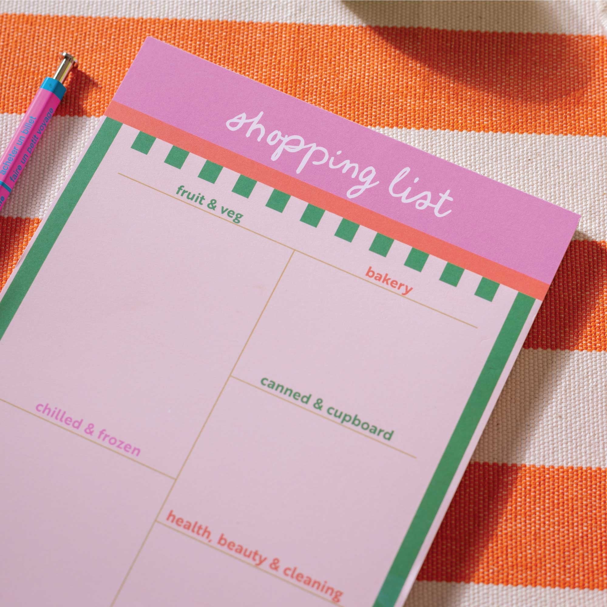Pink shopping list pad with a pen on an orange and white striped background.