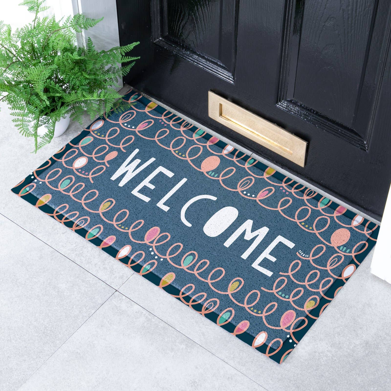 A rectangular doormat with a dark teal background, white 'WELCOME' text, and a colorful pattern of overlapping teardrop shapes, shown in front of a black front door on a light gray tiled floor with a green plant nearby.