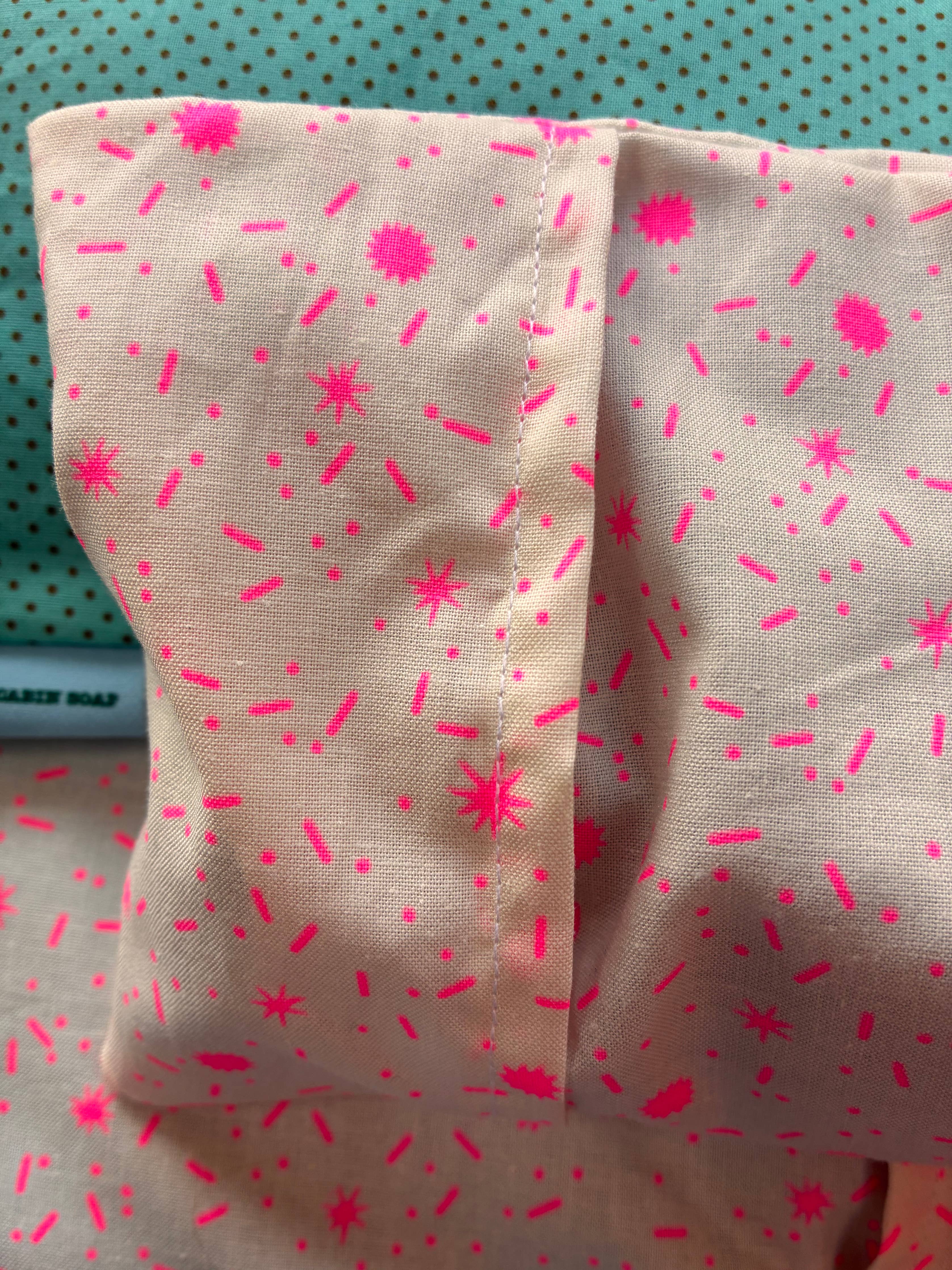 A close-up of a light beige fabric eye pillow, printed with a bright pink sprinkle pattern, held against a teal polka-dot background.