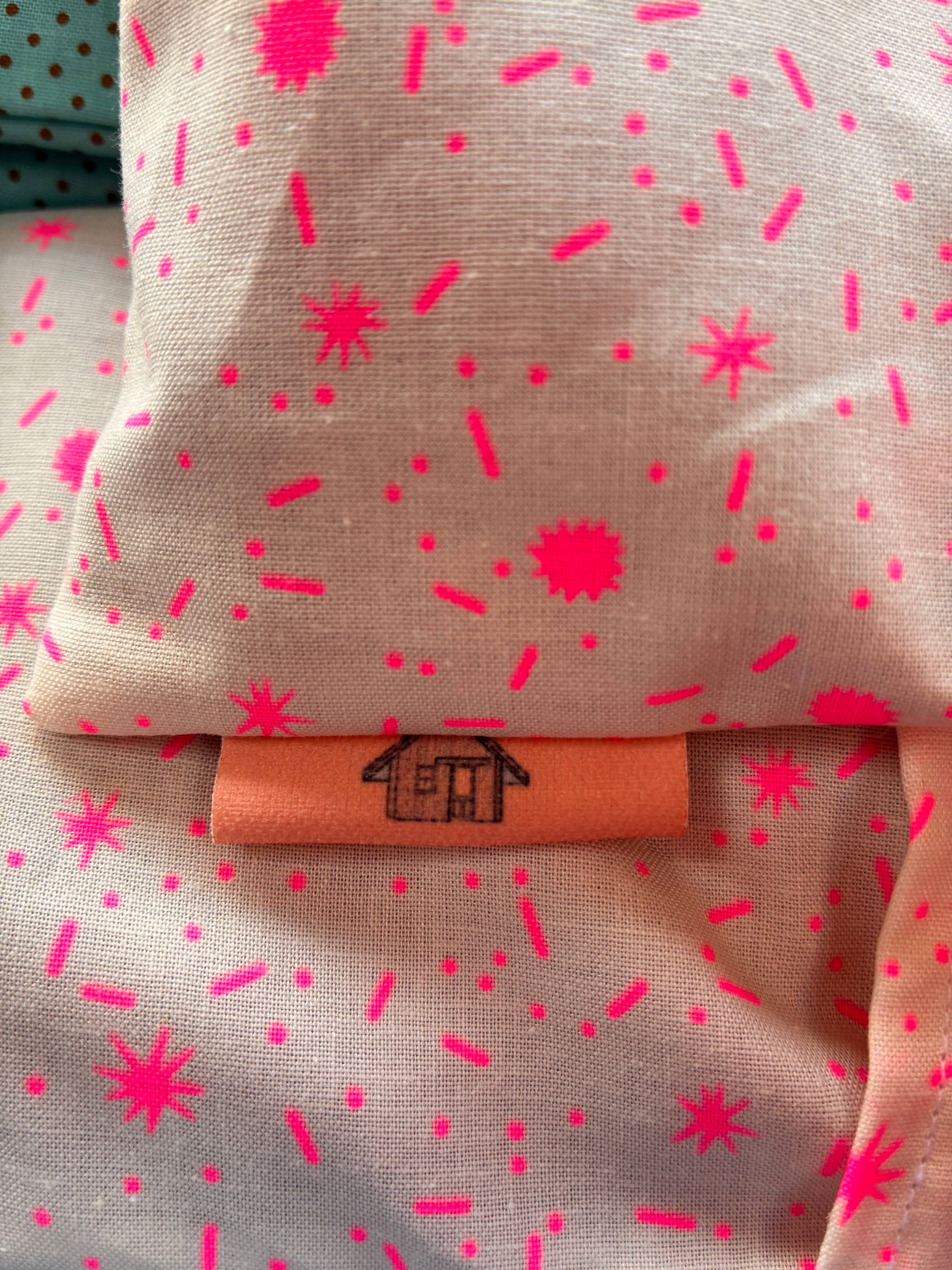 A close-up of an eye pillow, likely a pillow or cushion cover, printed with a vibrant hot pink confetti and starburst pattern. A peach-colored fabric label with a black hand-drawn house logo is sewn onto the front.