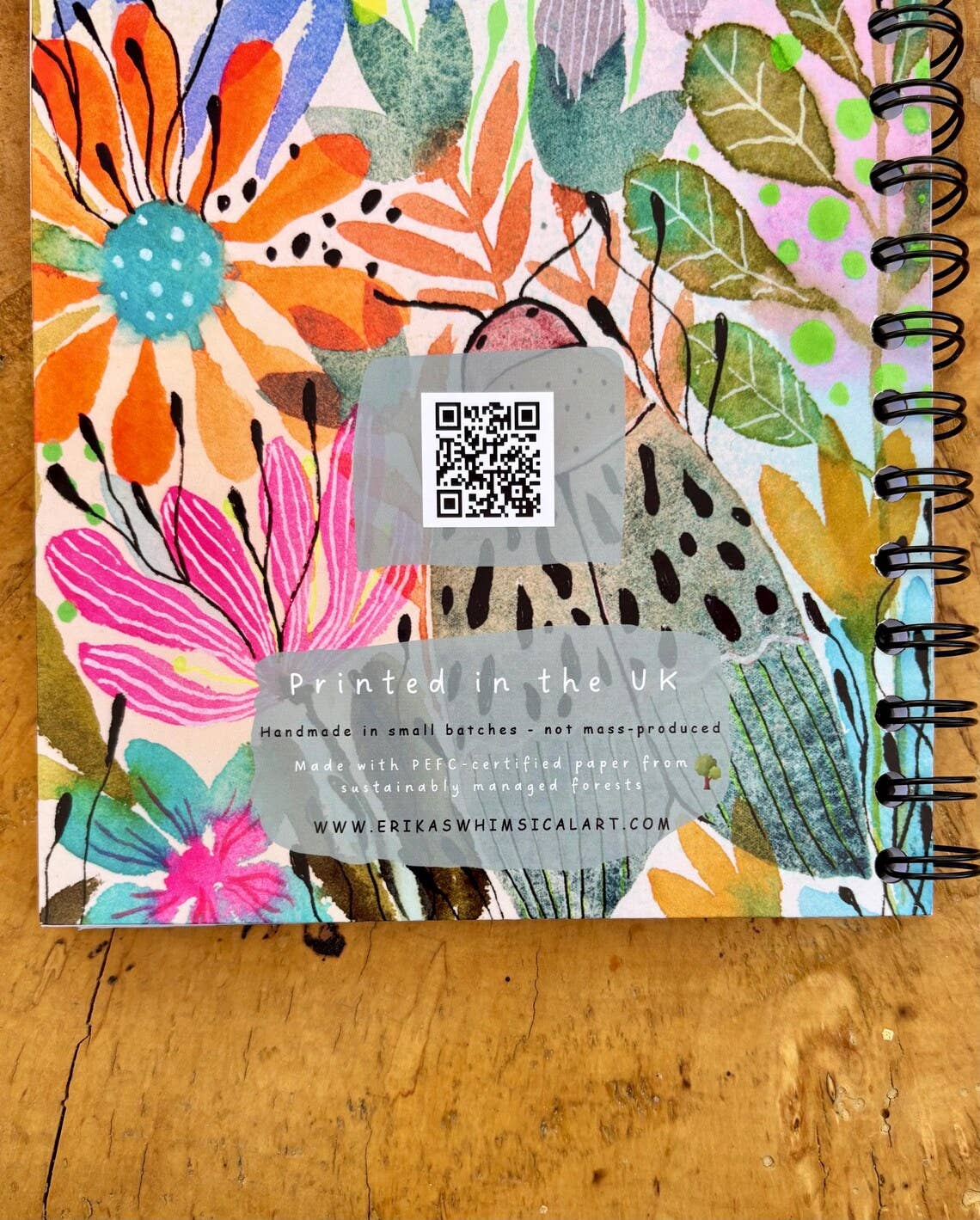 A spiral-bound notebook with a colorful floral and botanical pattern cover, photographed on a wooden surface. The cover features large illustrated flowers and leaves in orange, pink, teal, green, yellow, and purple, with a translucent grey sticker overlay containing a QR code and text that reads 'Printed in the UK'.