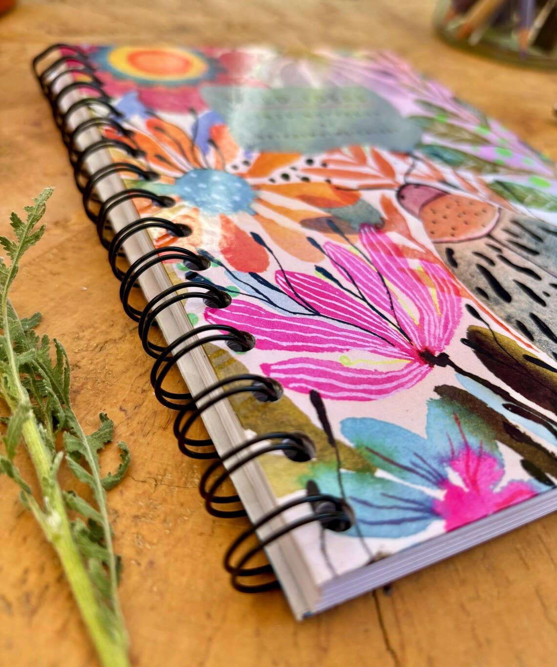 A spiral-bound notebook with a colourful floral and botanical illustration on the cover, placed on a wooden surface with a green stem in the foreground.