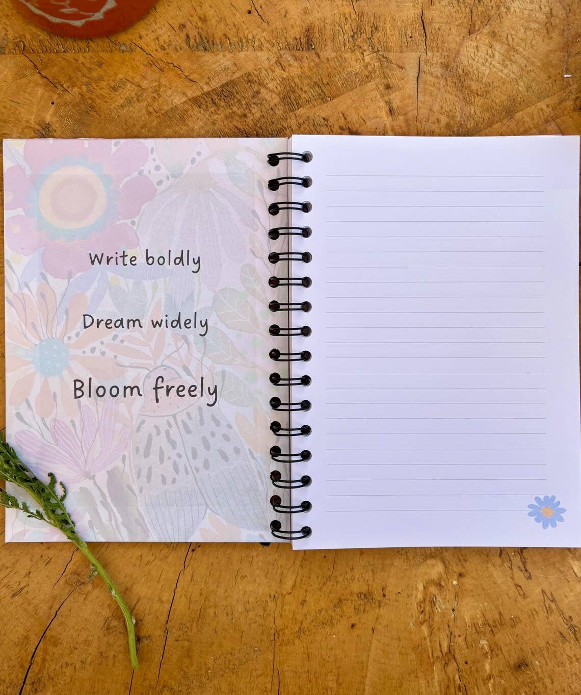An open spiral-bound journal lying on a wooden surface, with a pastel floral cover and lined white pages. Handwritten text on the left page reads 'Write boldly, Dream widely, Bloom freely,' and a small blue flower illustration appears in the bottom right corner.