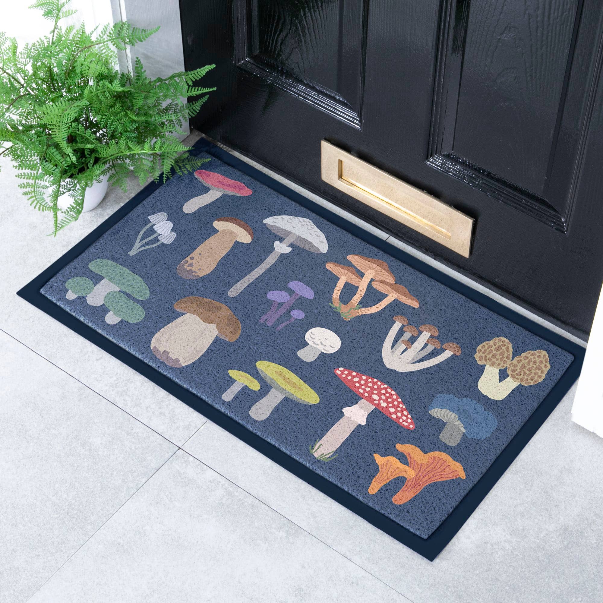 A rectangular doormat with a dark blue-gray background and colorful illustrated mushrooms, placed in front of a black front door on a light tile floor with a potted fern nearby.