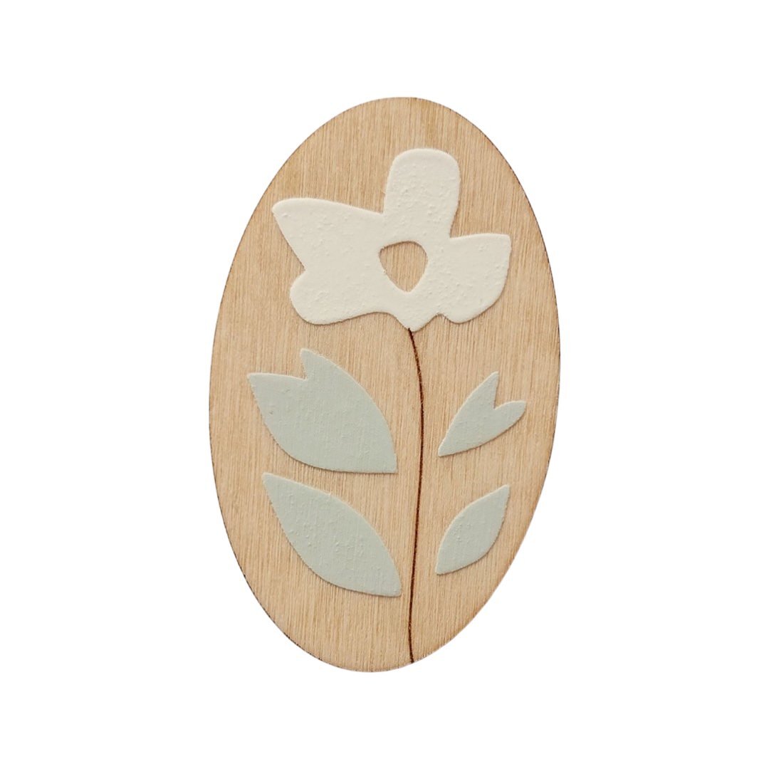 An oval wooden brooch with a white flower, brown stem and green leaves.