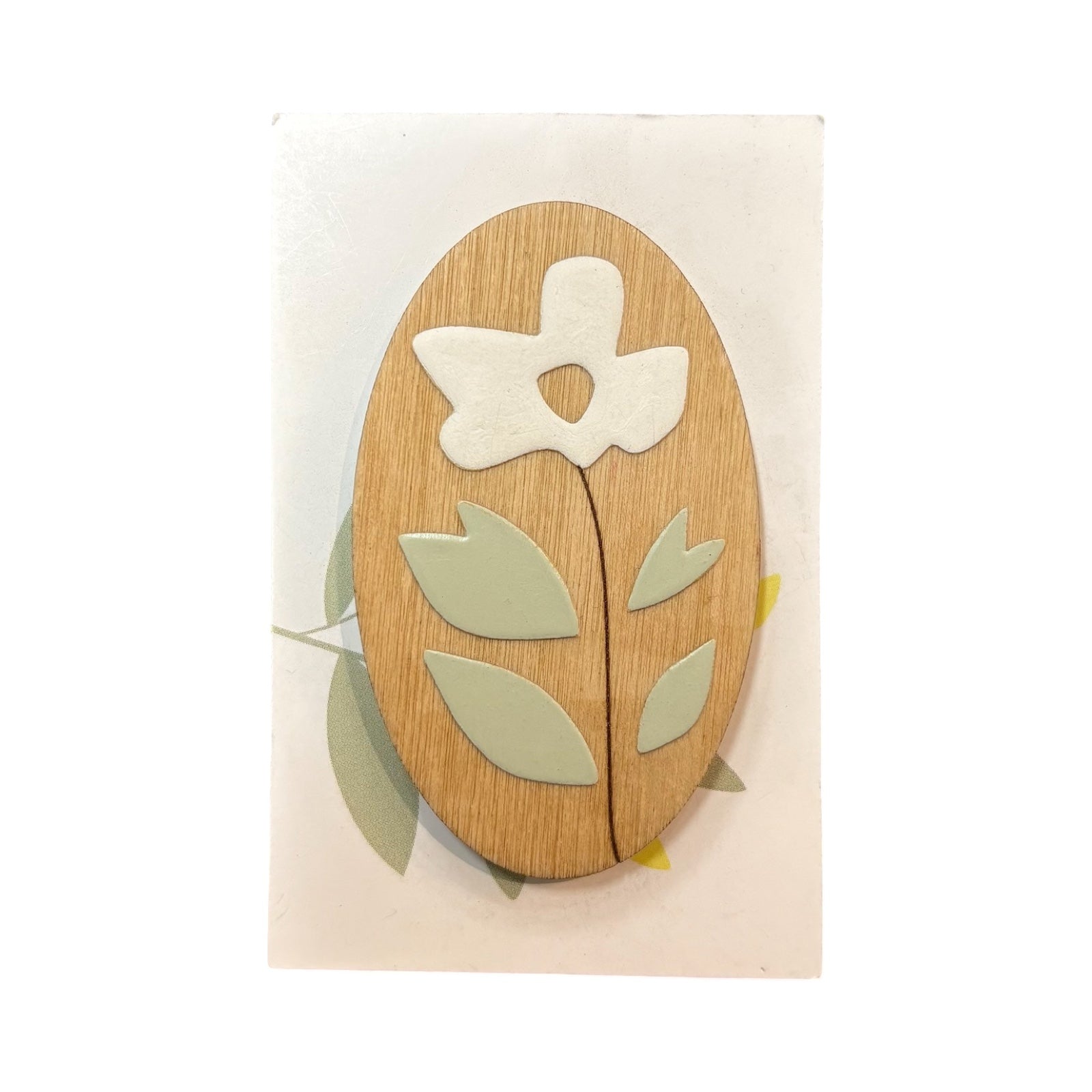 An oval wooden brooch with a white flower, brown stem and green leaves on a white backing card.